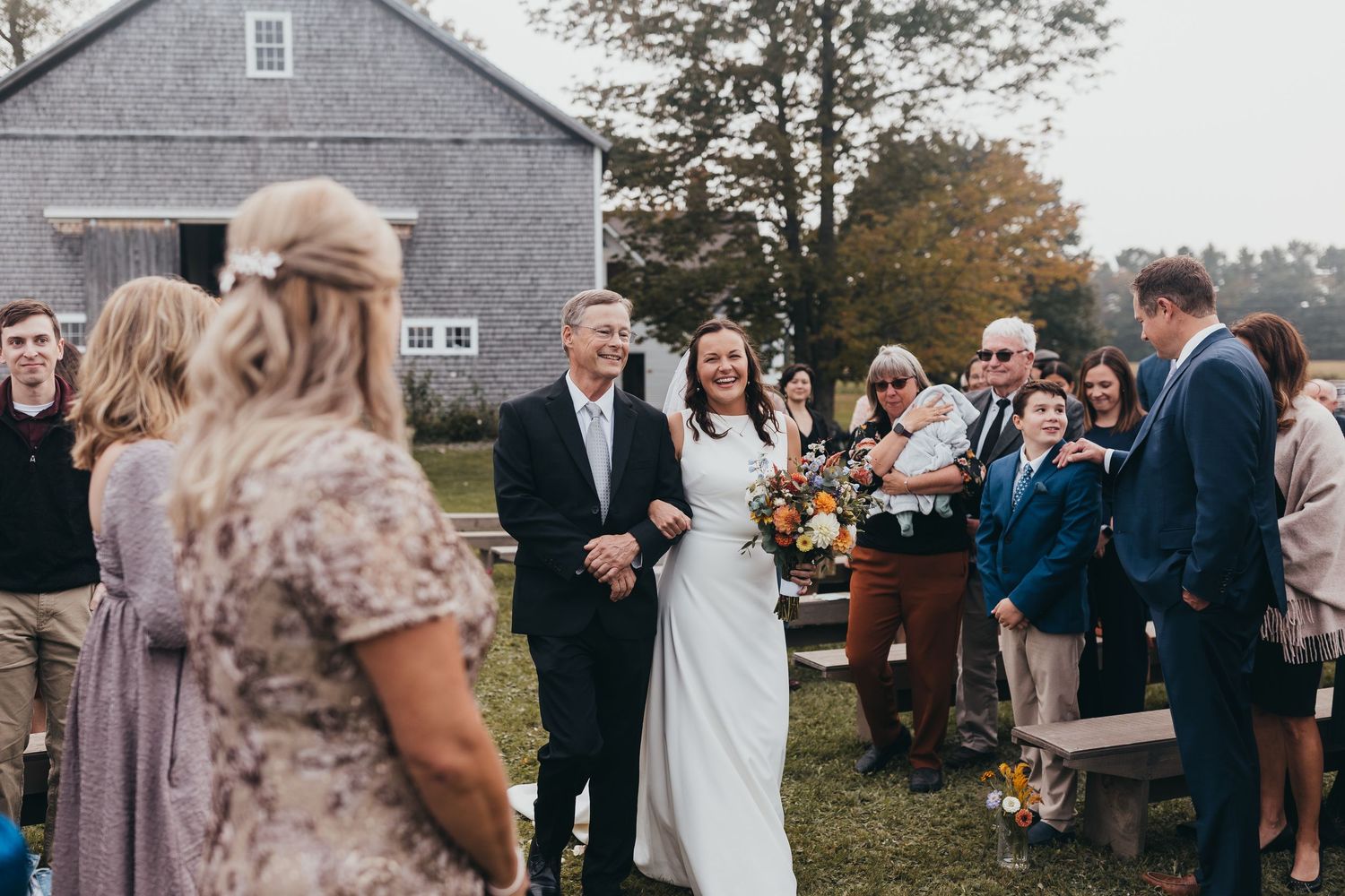 Wedding ceremony guests gathered outside a grey building during an outdoor celebration.
