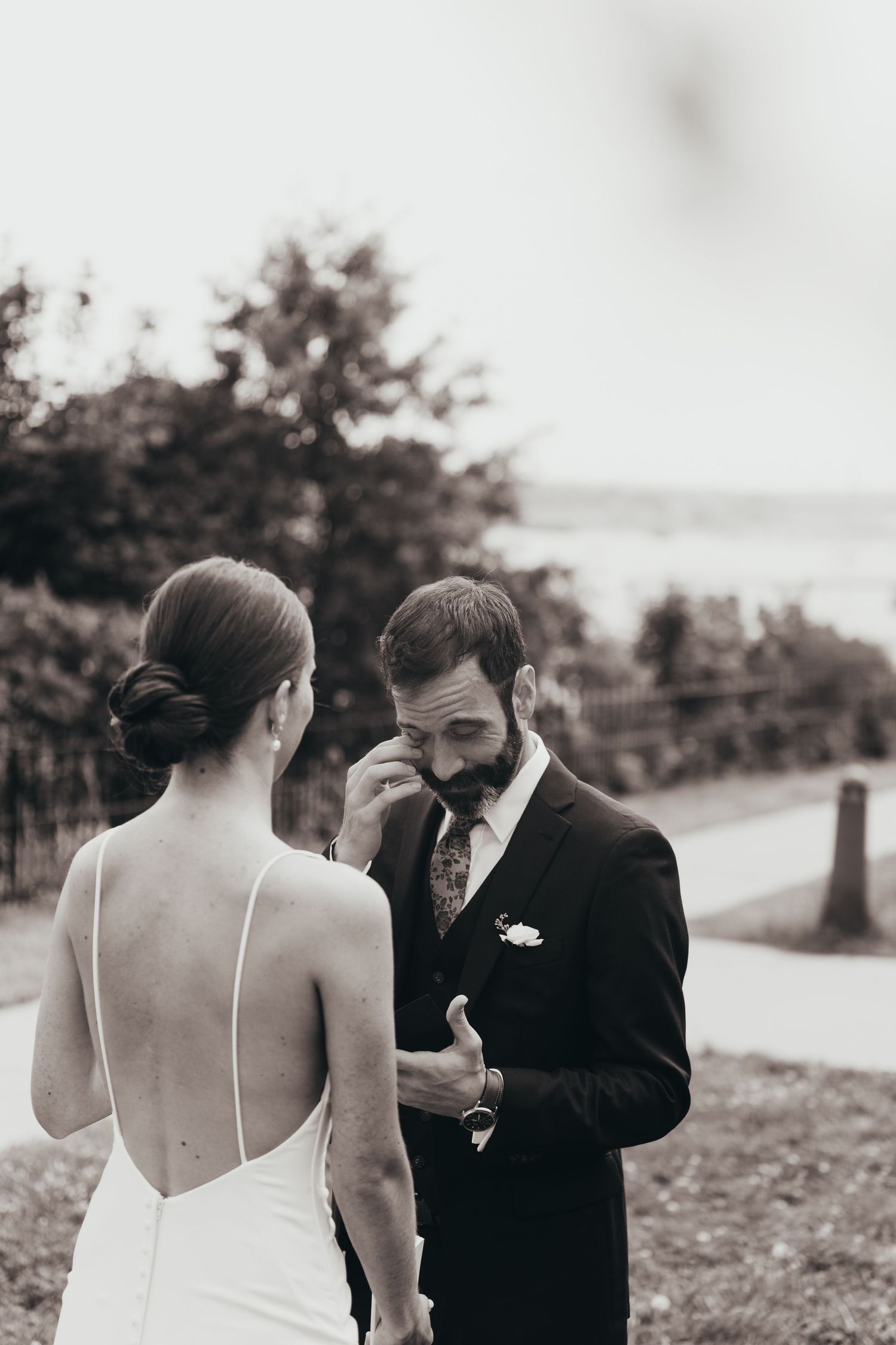 Black and white photo of emotional groom wiping tears as bride in backless gown stands before him at outdoor wedding.