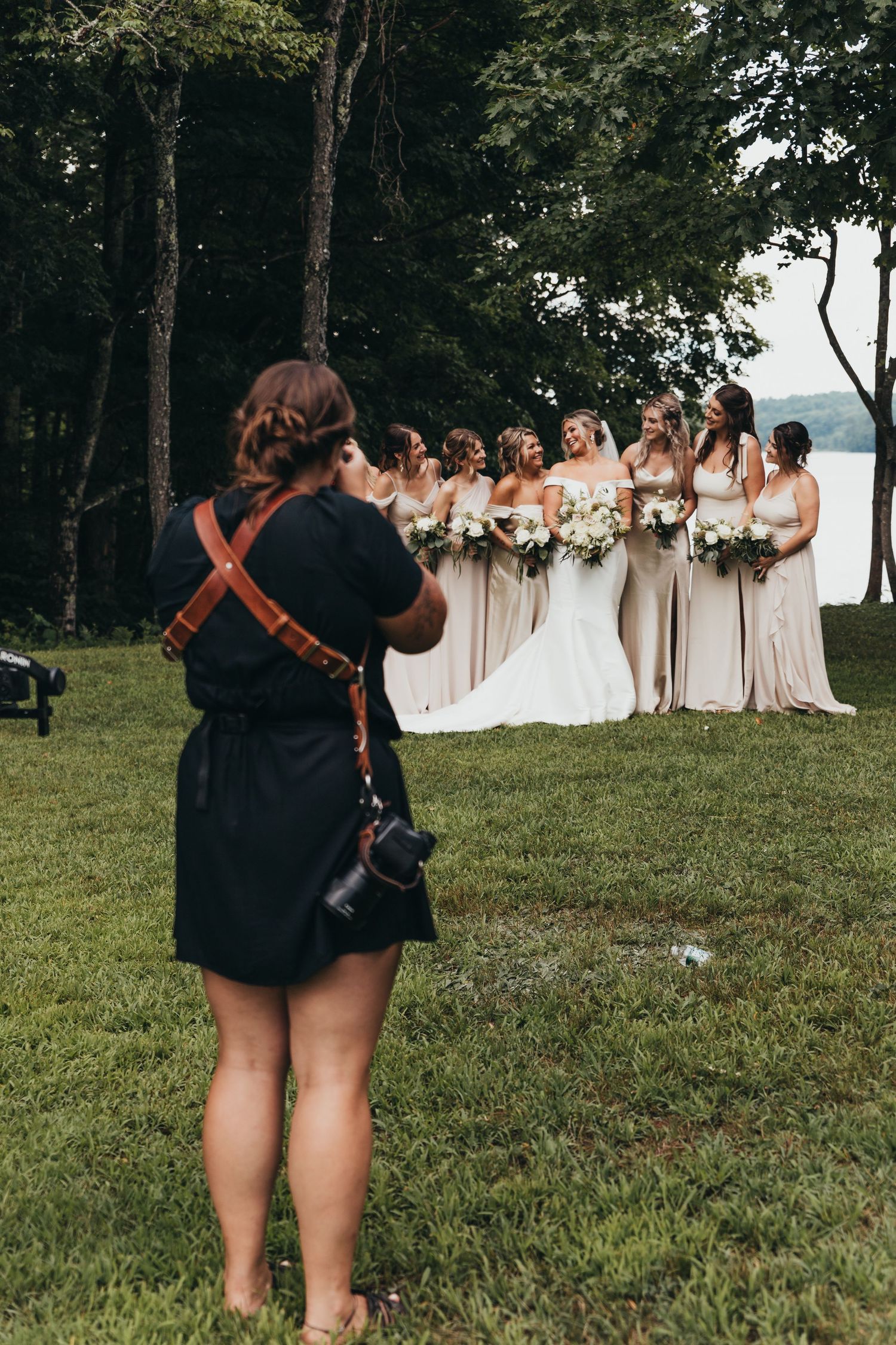 Wedding photographer capturing bridesmaids in light dresses on grassy lawn.