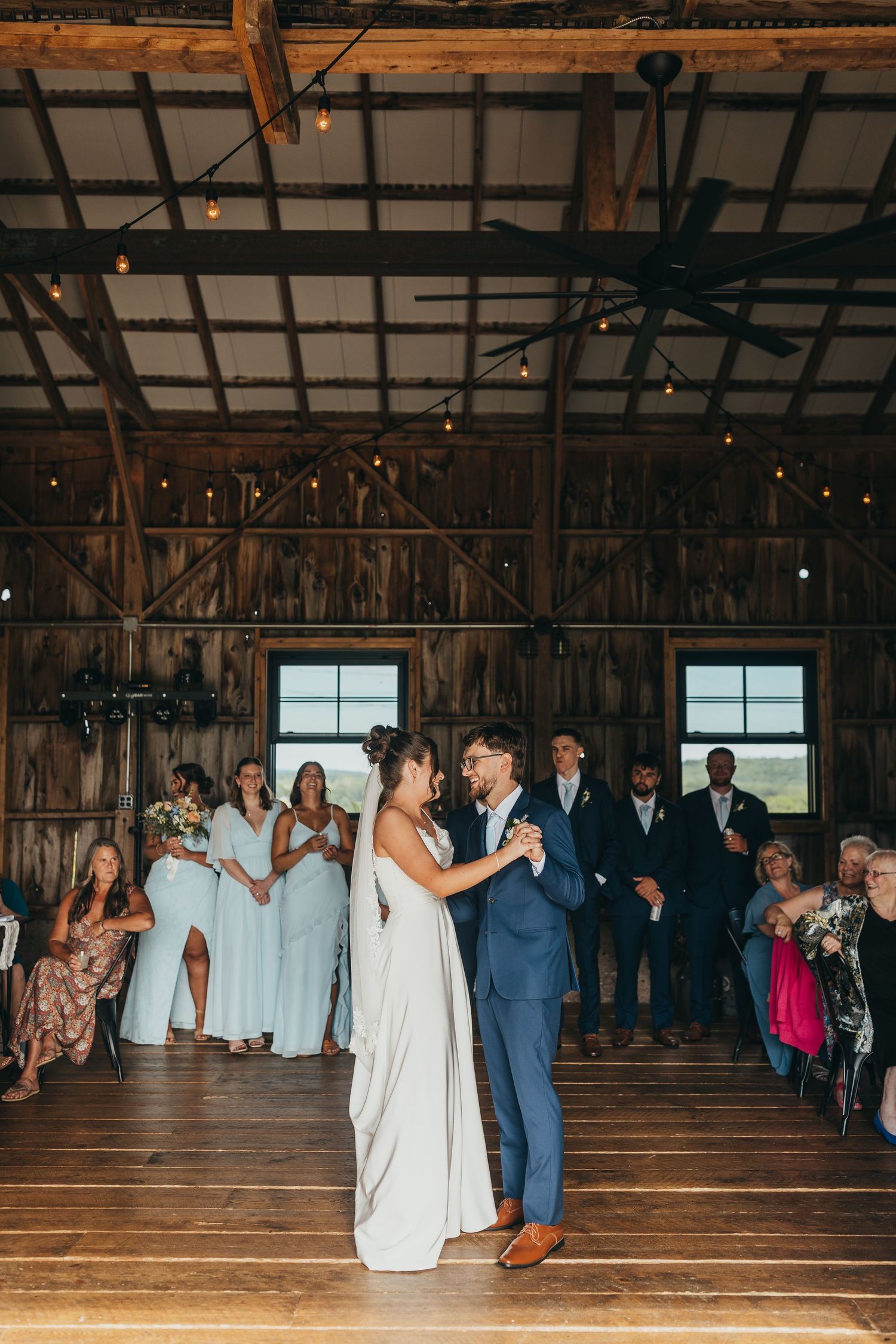 Bride and groom share first dance in rustic barn venue with string lights, bridesmaids, and guests watching.