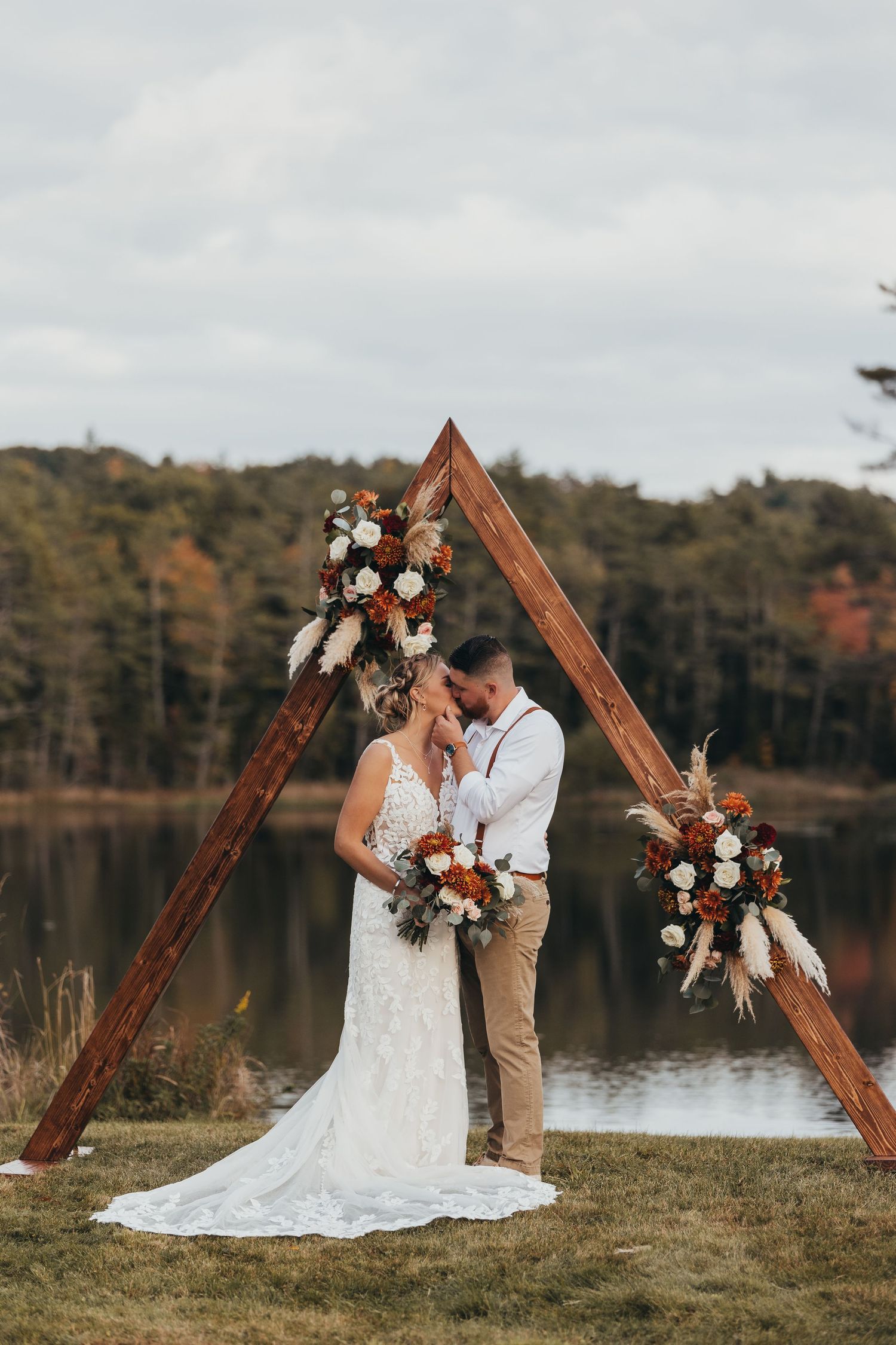 Wedding couple posing by wooden triangle arch with floral decorations near lakeside.