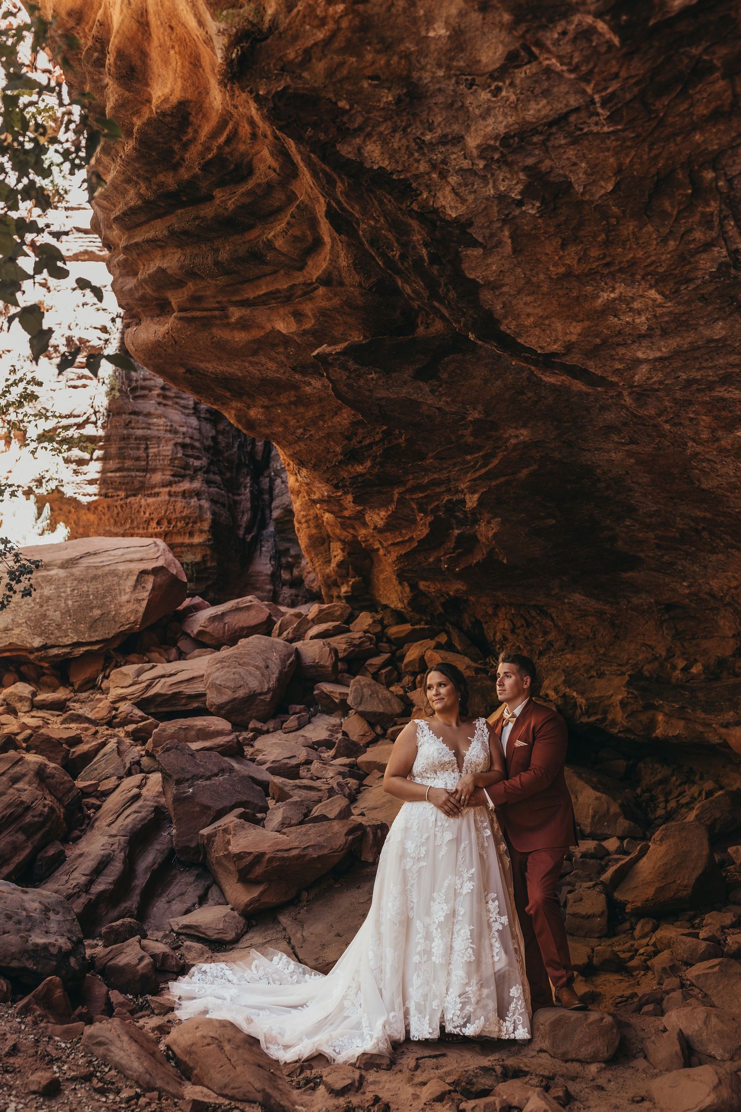 Bride and groom pose romantically beneath a red rock cave overhang, surrounded by rustic boulders in a stunning outdoor setting.