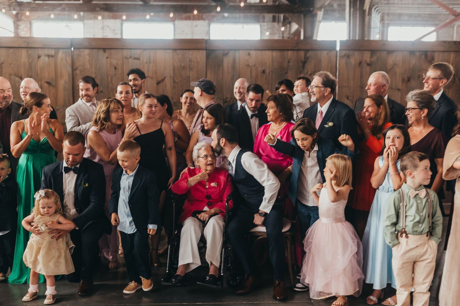Large group of wedding guests celebrating and dancing in a rustic indoor venue.
