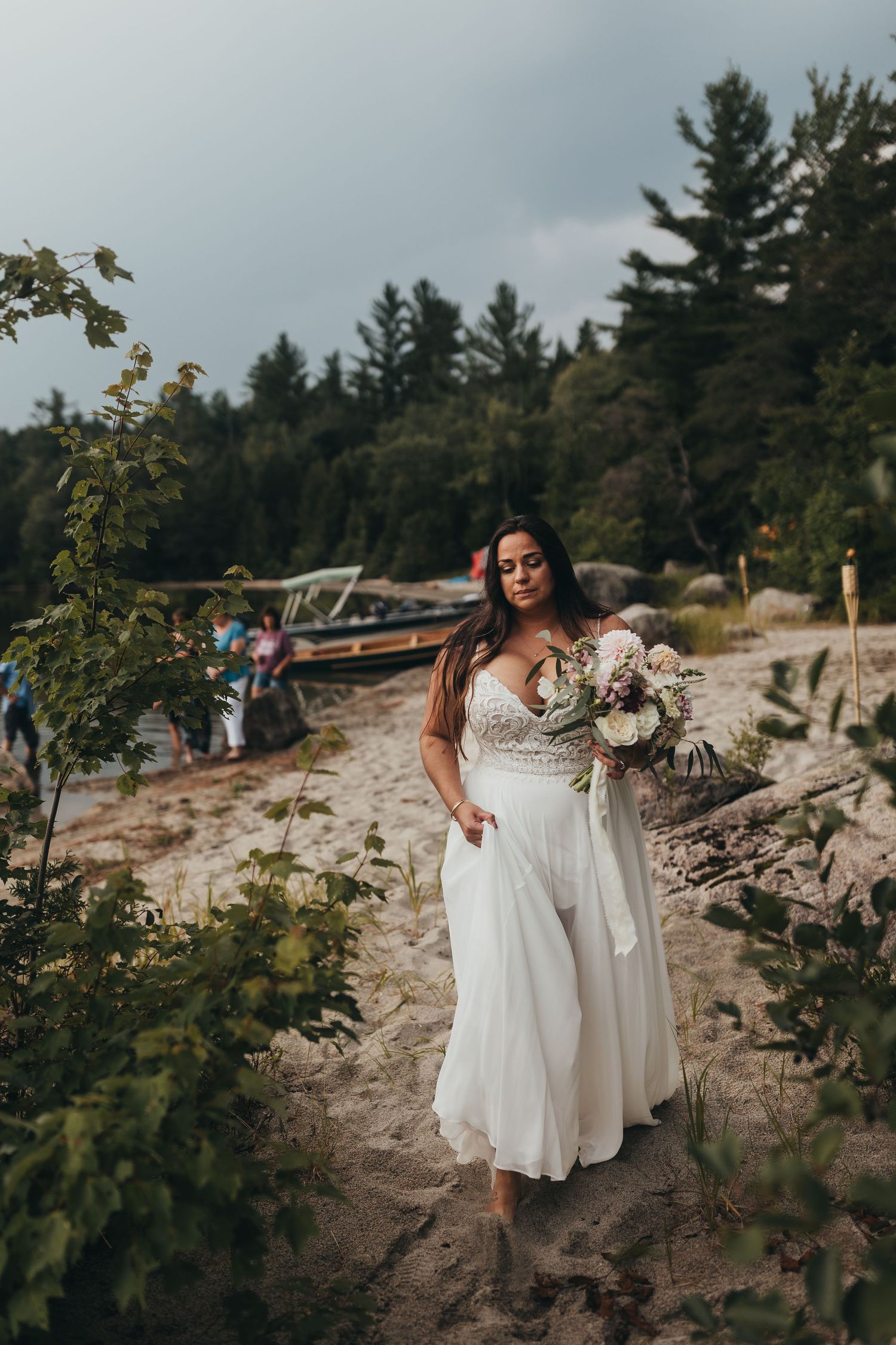 Bride in white gown holding bouquet walks barefoot on sandy beach path with trees and boat in background.