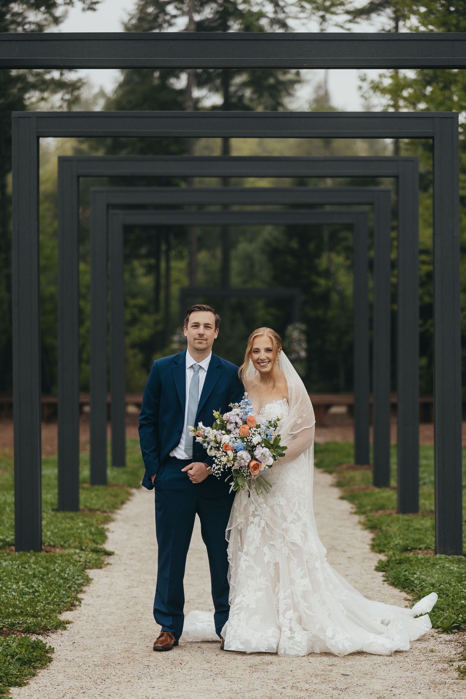 A couple walks along a path framed by dark architectural pillars in an outdoor wedding setting.