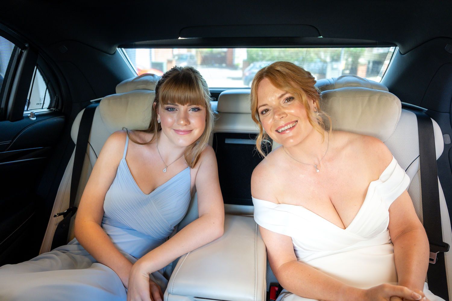 Bride in white gown and bridesmaid in blue dress smiling in the back of a wedding car.