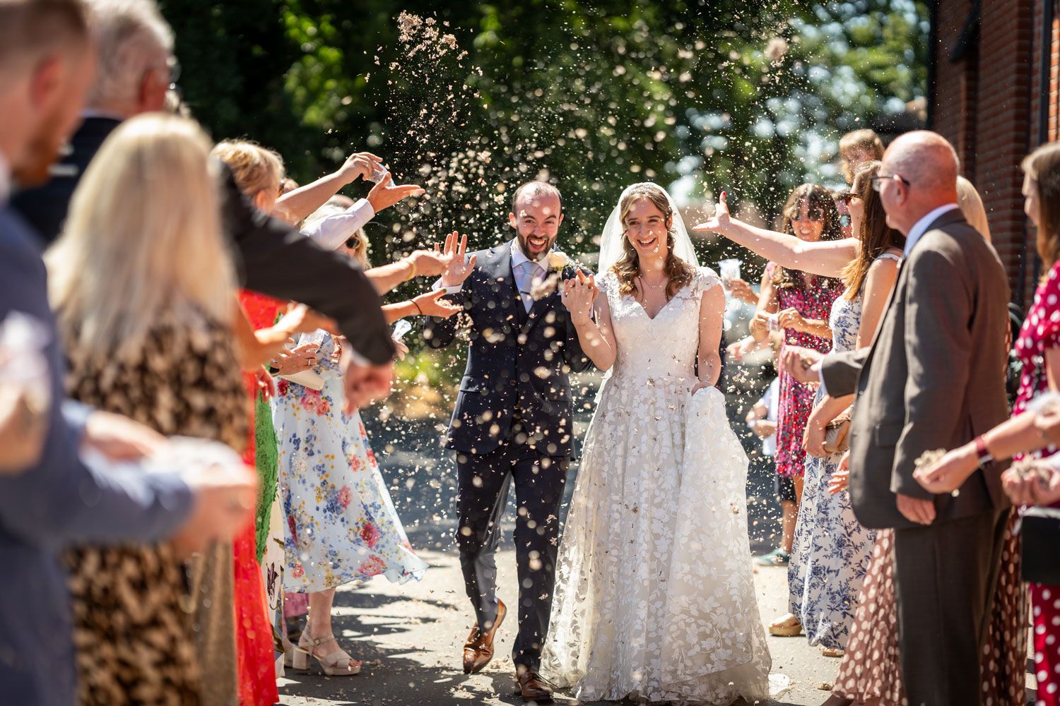 Bride and groom walk through a joyful confetti shower thrown by smiling wedding guests outside.