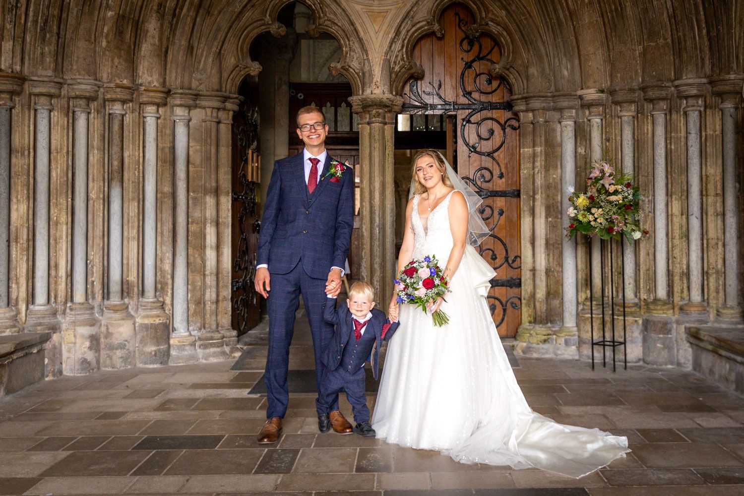 Bride and groom with young son posing in the ornate stone archway of a historic cathedral.