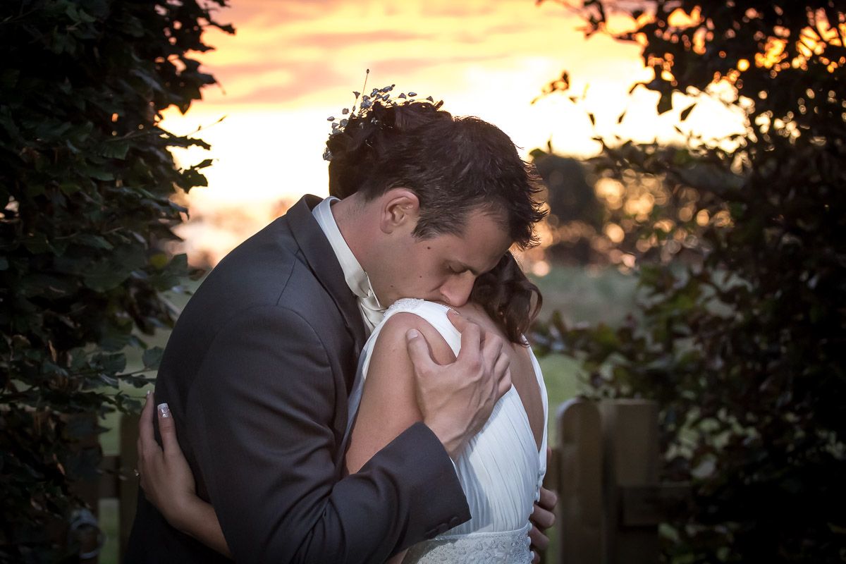 A tender intimate moment between a couple in formal attire embracing at sunset with lush foliage framing the scene.