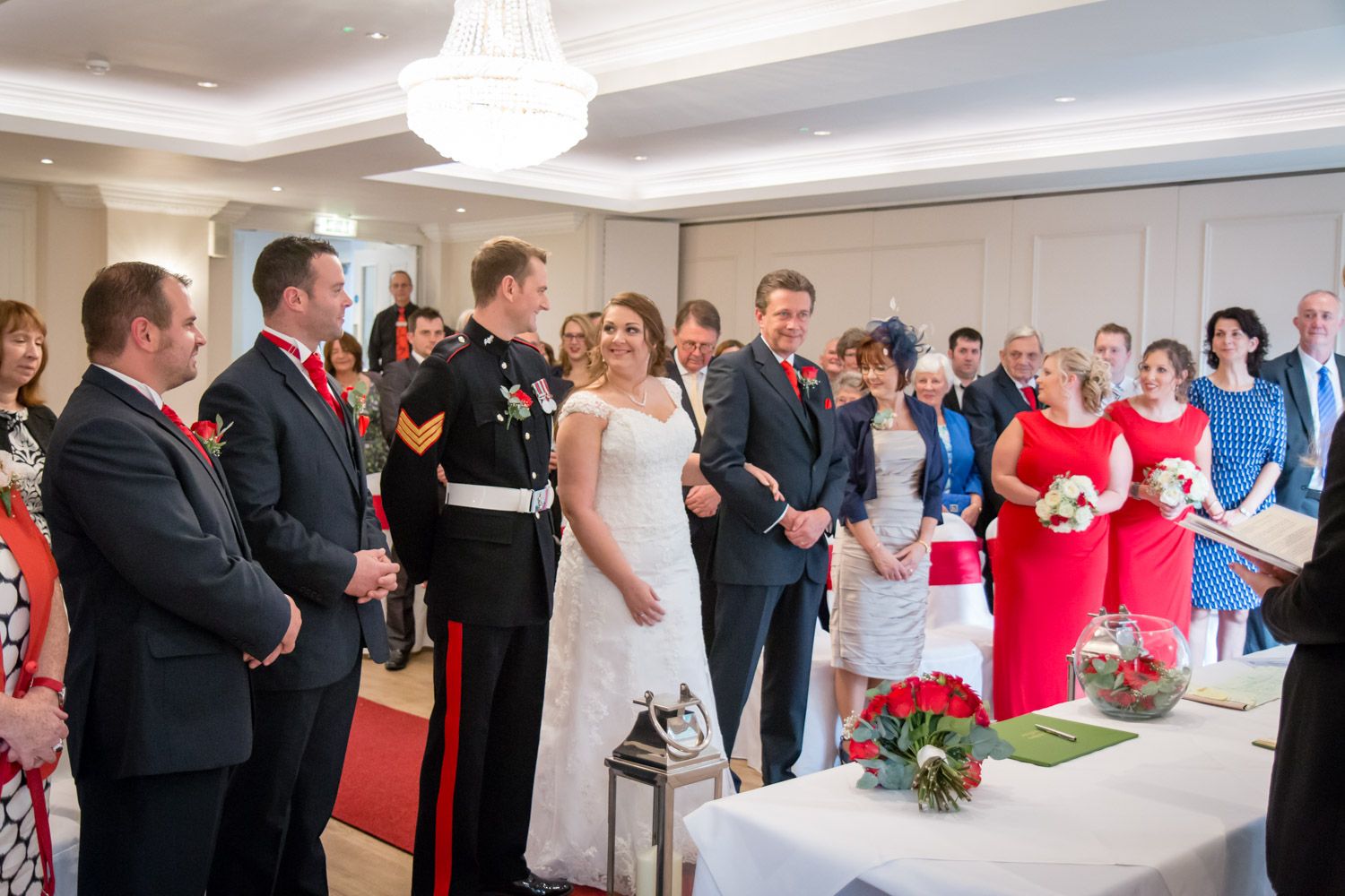 Wedding ceremony guests gather around a signing table with elegant decor and a crystal chandelier overhead.