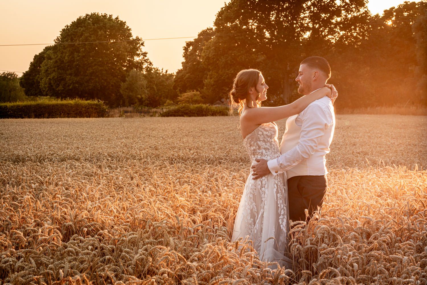Bride and groom embracing romantically in a golden wheat field at sunset.