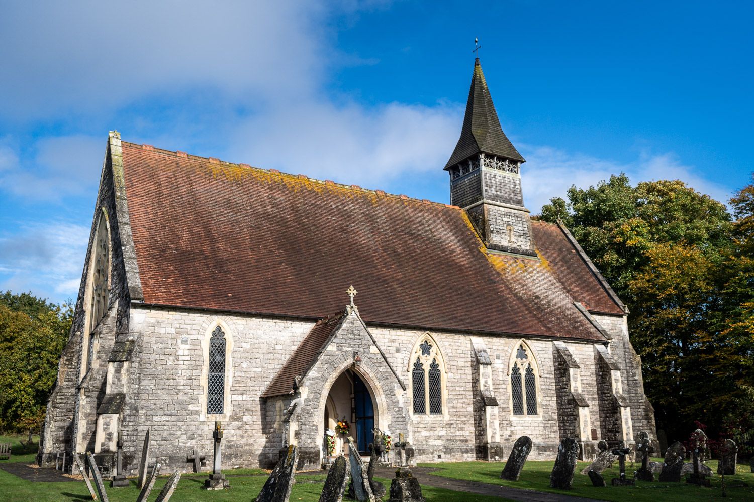 Small stone church with pointed spire and brown roof situated among trees against blue sky on sunny day.