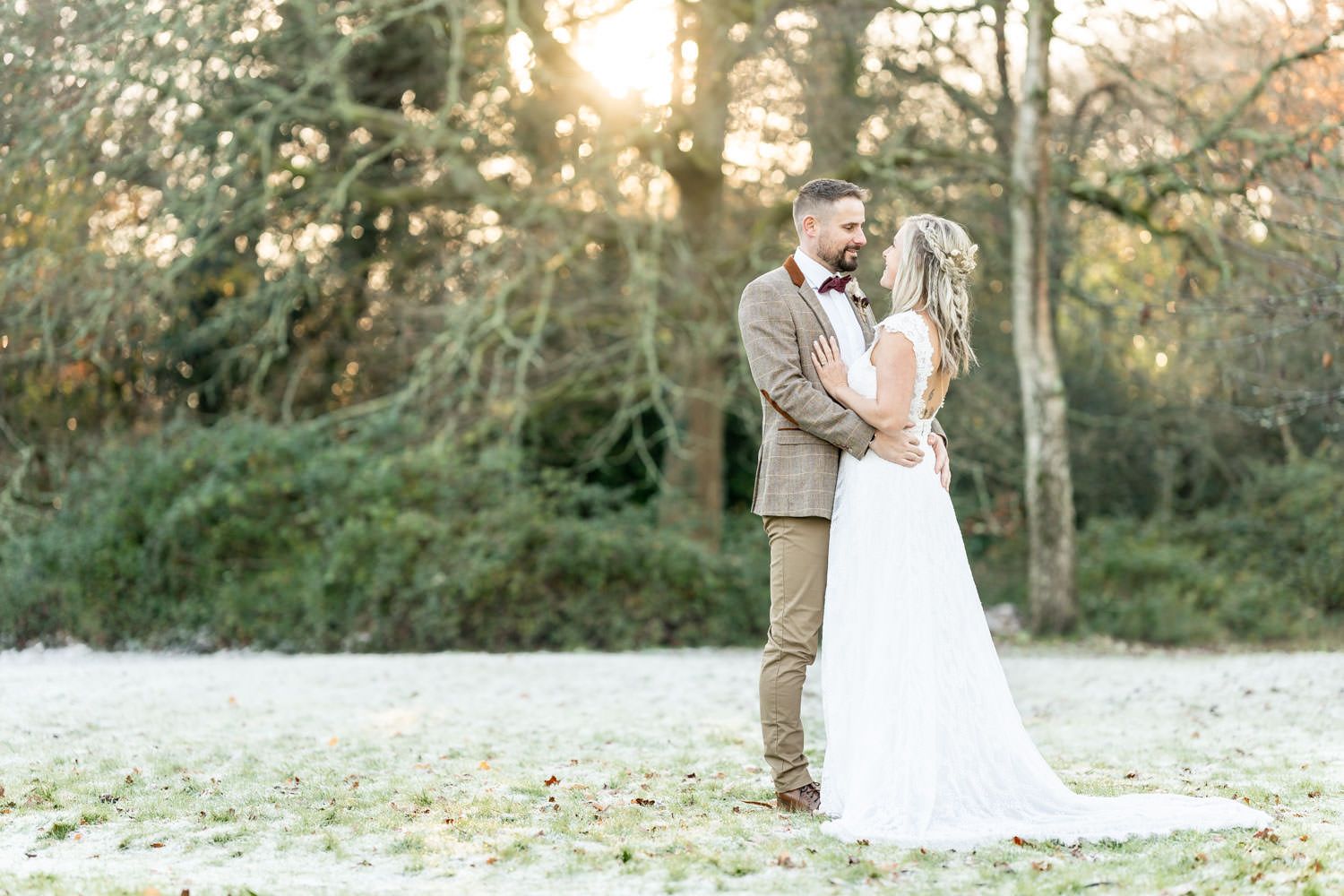 A couple in wedding attire embrace outdoors at sunset on a frosty ground with trees in the background.