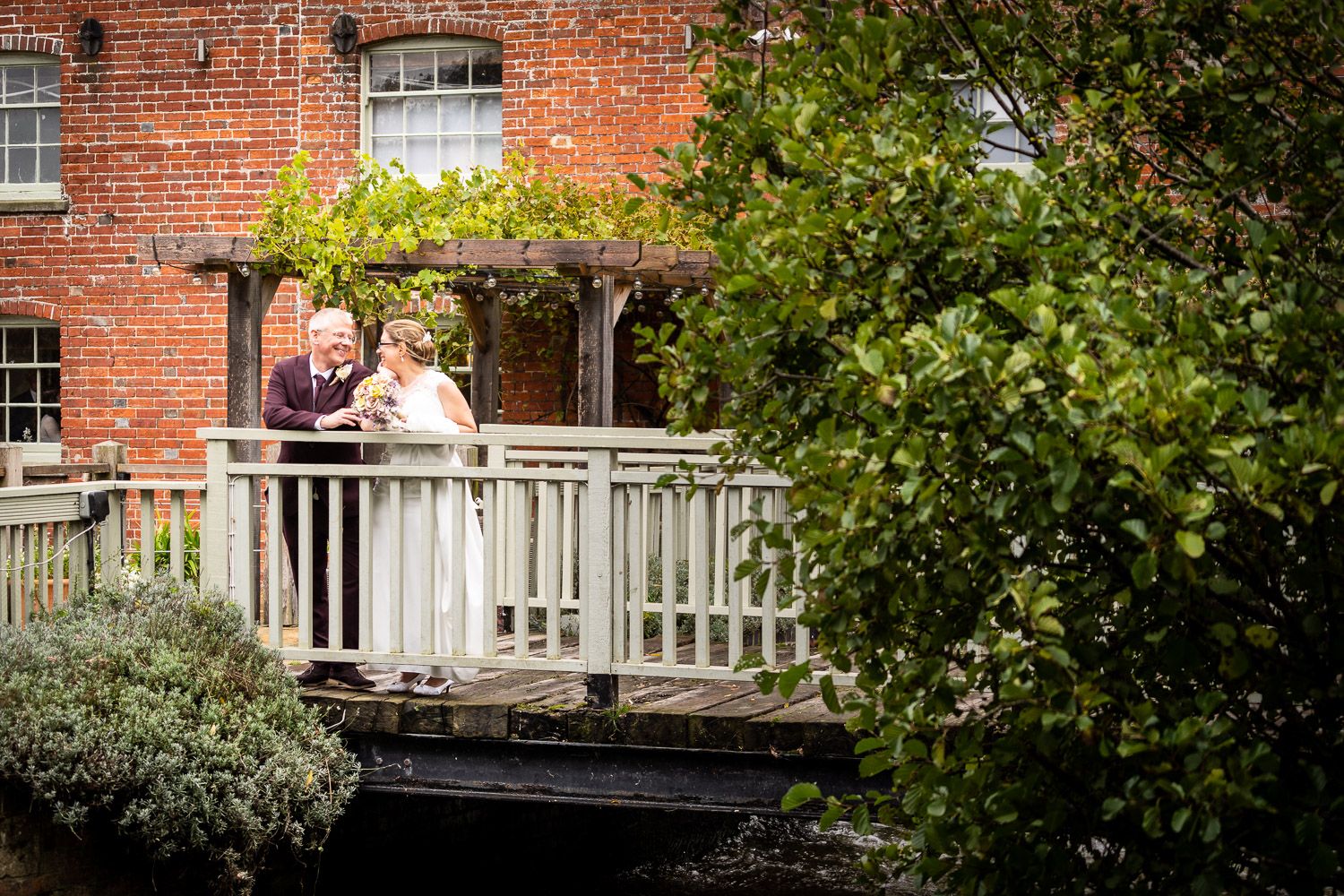 Wedding couple stands on a wooden bridge deck by a historic red brick building surrounded by lush greenery.
