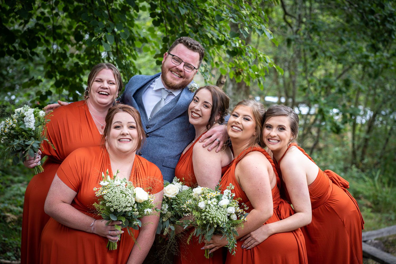 Wedding party with bridesmaids in bright coral dresses holding white flower bouquets pose together outdoors in nature.