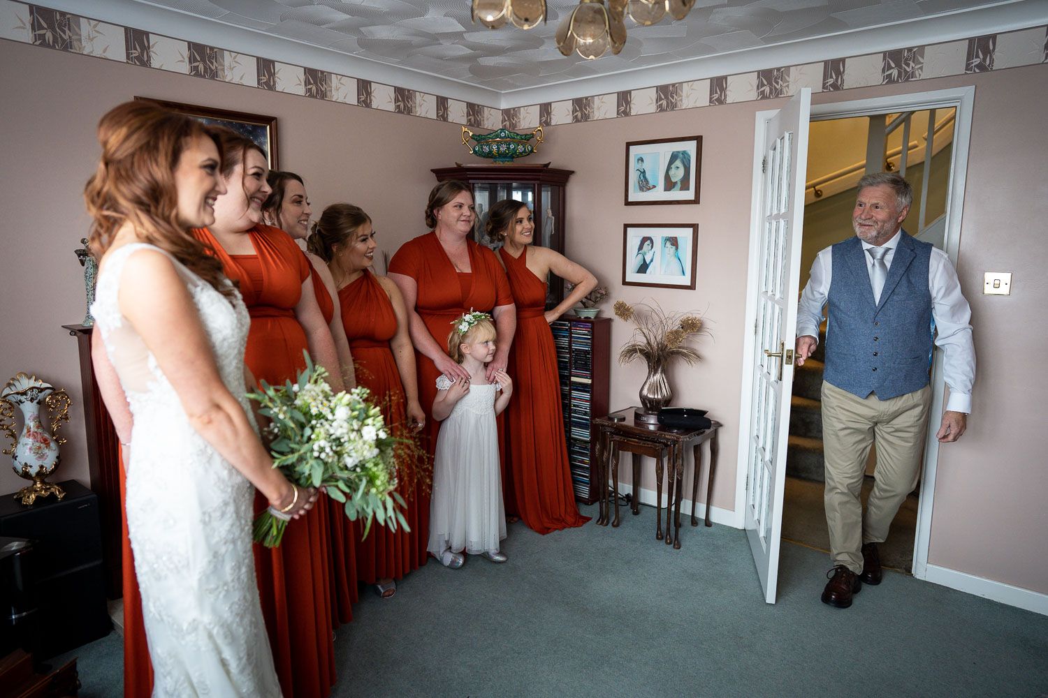 Wedding party in rustic orange bridesmaid dresses and white bridal gown await in room as someone enters through doorway.