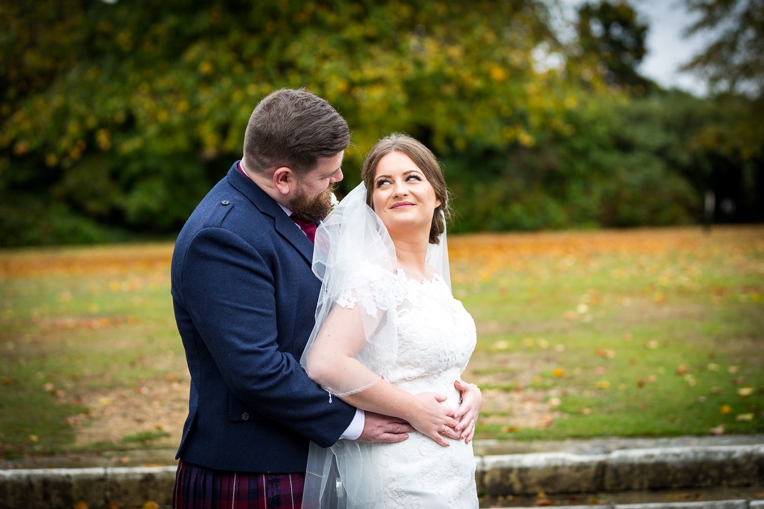 A couple in wedding attire embraces in a park with autumn foliage in the background.