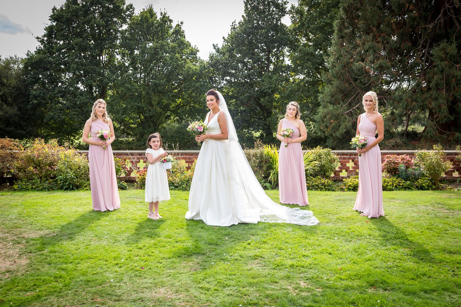Wedding party in light pink bridesmaid dresses and white wedding gown pose on manicured lawn with garden backdrop.