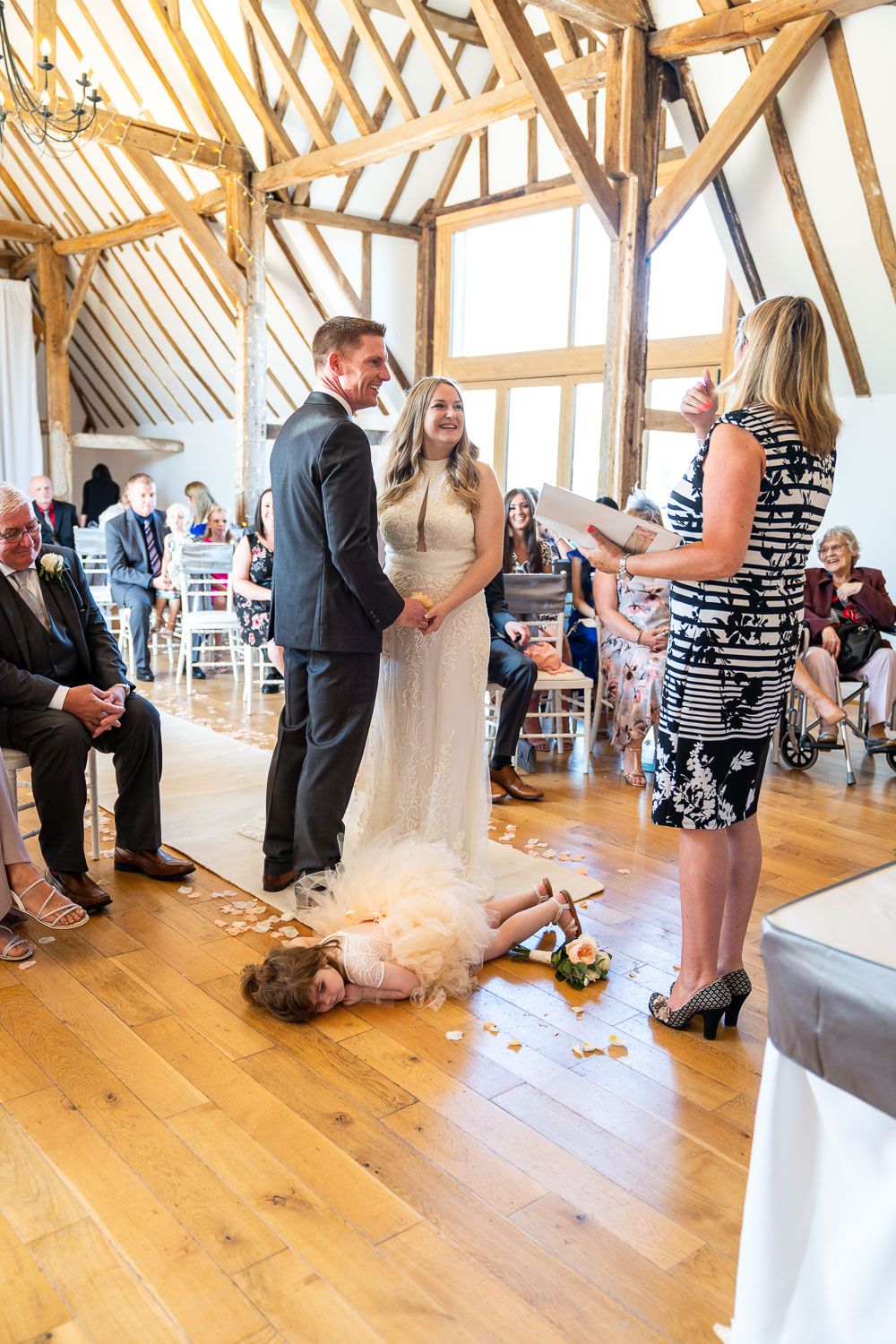 Wedding ceremony in a rustic wooden beam venue with a young child playfully rolling on the hardwood floor.