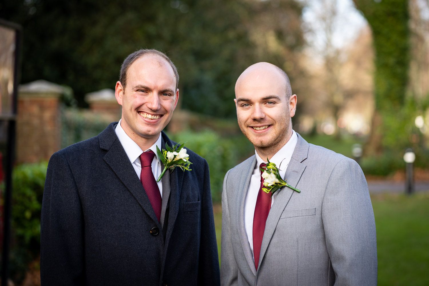 Two men in formal attire with matching burgundy ties and boutonnieres smile at an outdoor wedding.