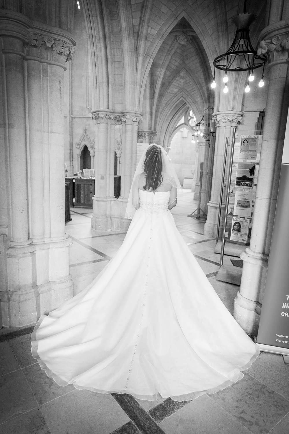 A bride in a flowing white wedding gown with long train stands in a gothic cathedral with arched stone columns and vaulted ceilings.