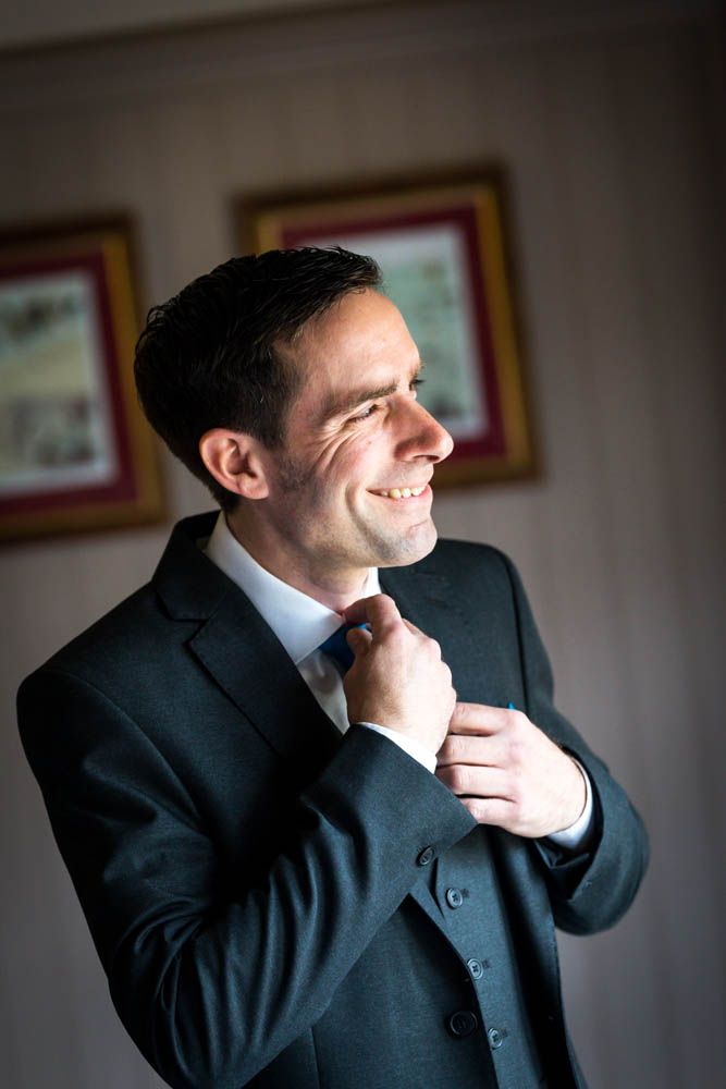 A person in a dark suit and tie adjusts their collar while smiling in a softly lit room with framed artwork on the wall.