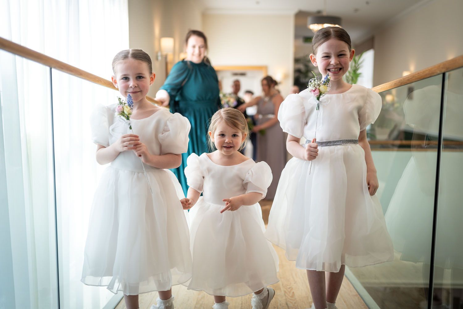 Three excited young attendants in white dresses hold their bouquets while walking down a hallway.