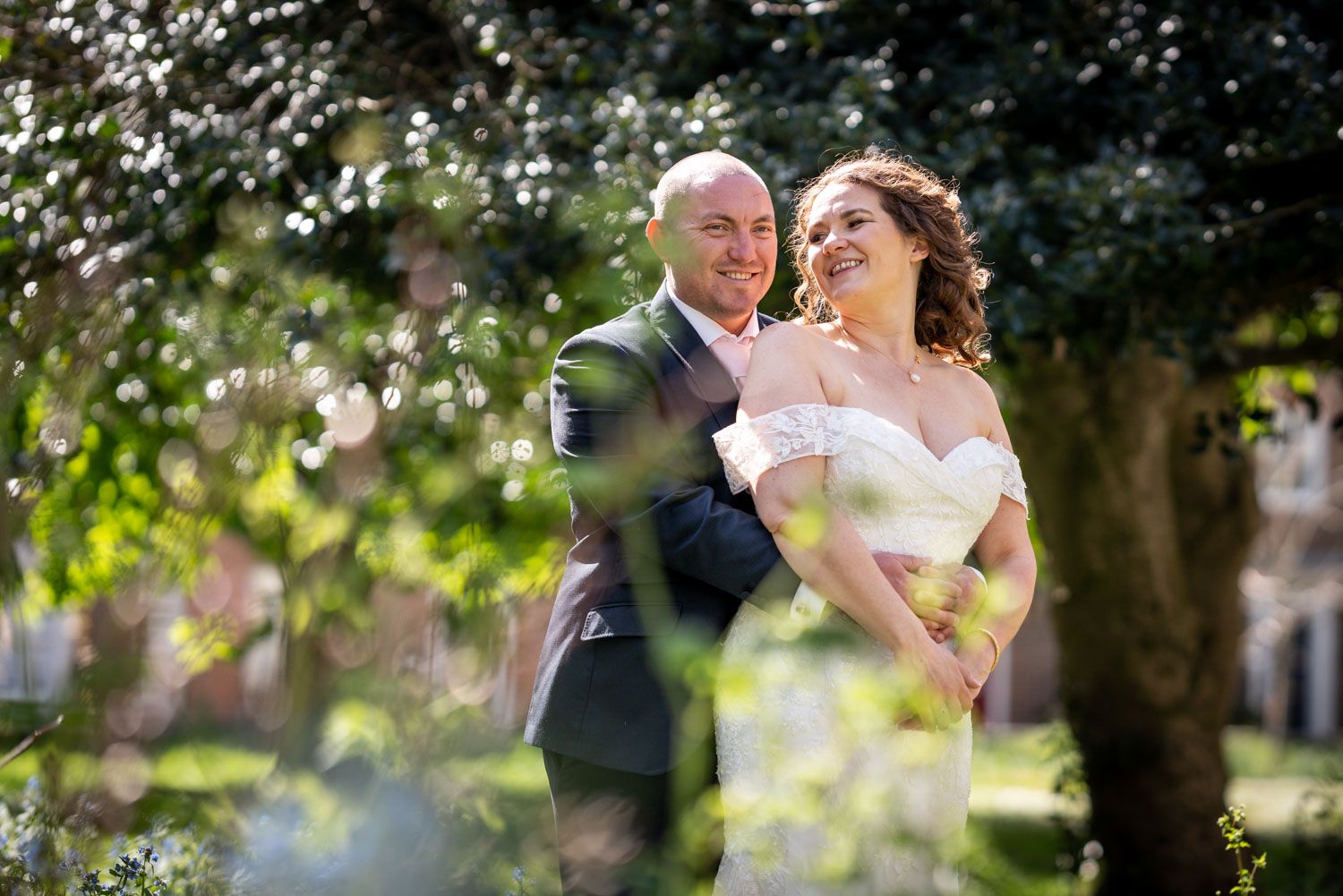 Smiling couple in wedding attire pose together in sunlit garden surrounded by sparkling bubbles.
