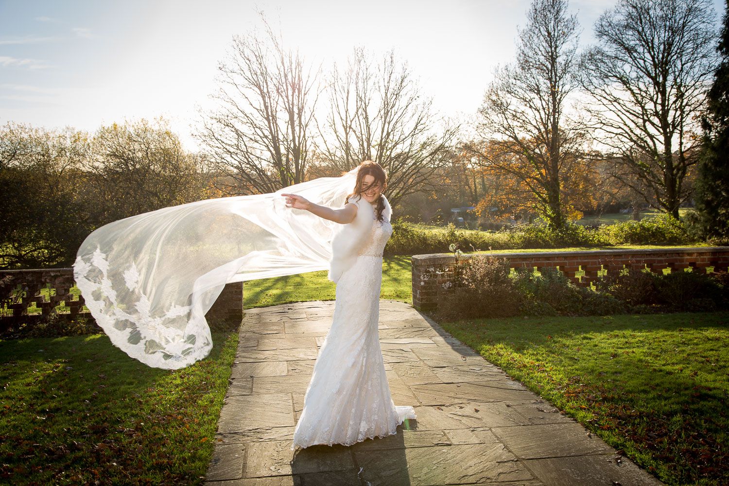 A bride's veil gracefully flows in the wind on a stone pathway surrounded by sunlit garden grounds and bare trees.