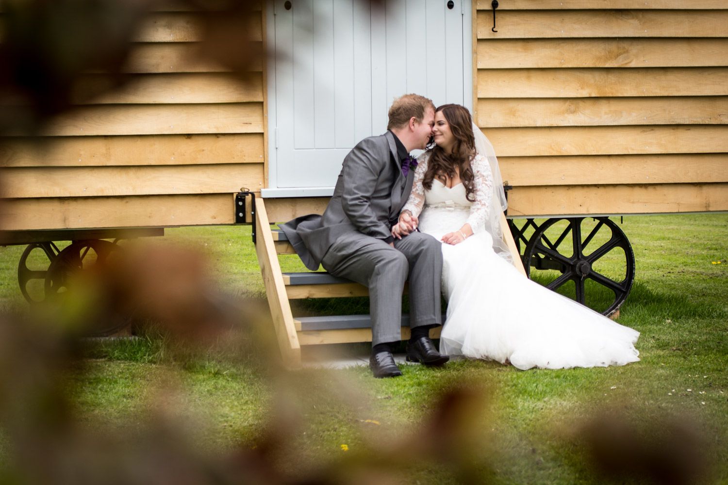 A couple in wedding attire sits close together on rustic wooden steps by a barn on their special day.