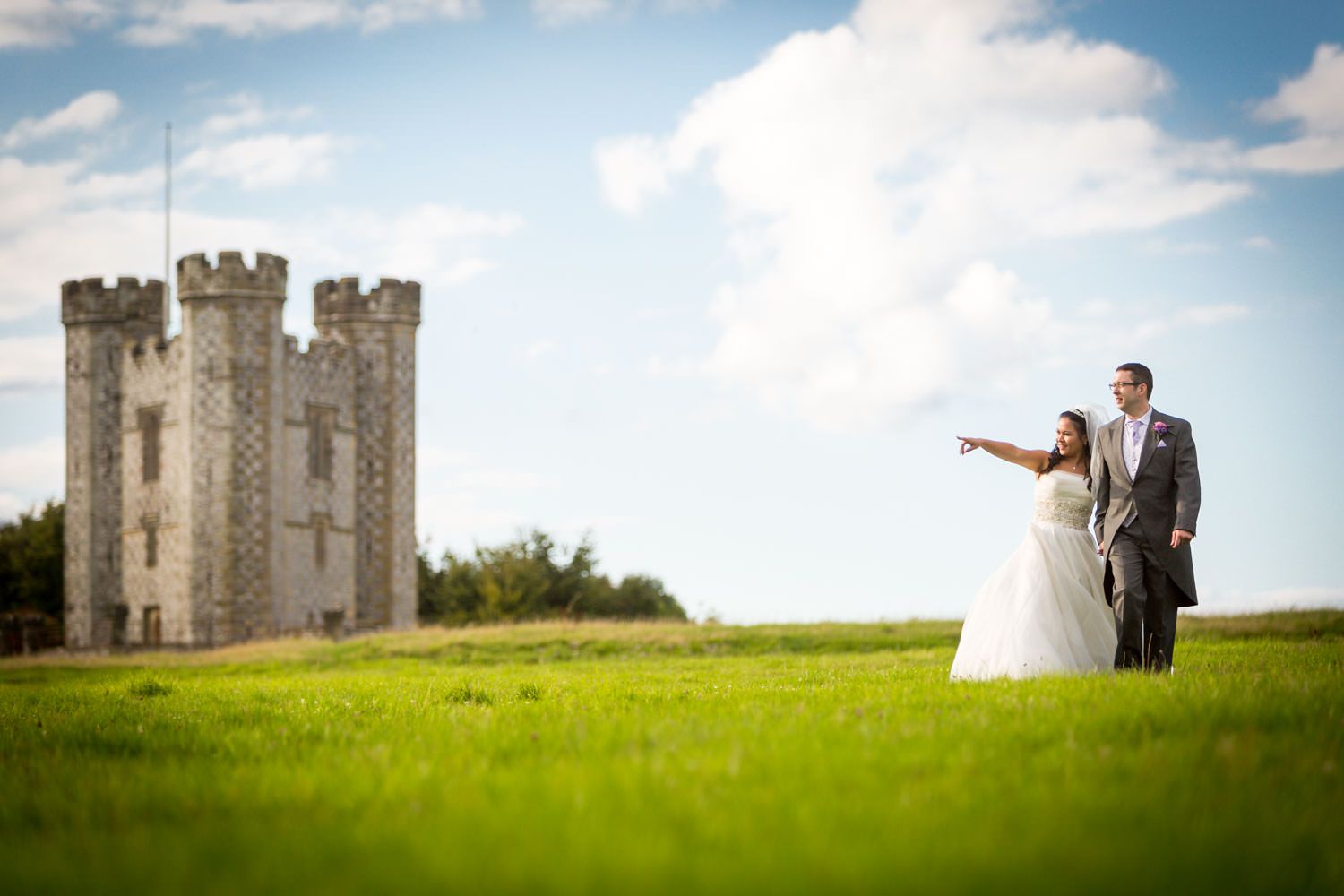 Bride and groom stand on green field with medieval castle tower backdrop.