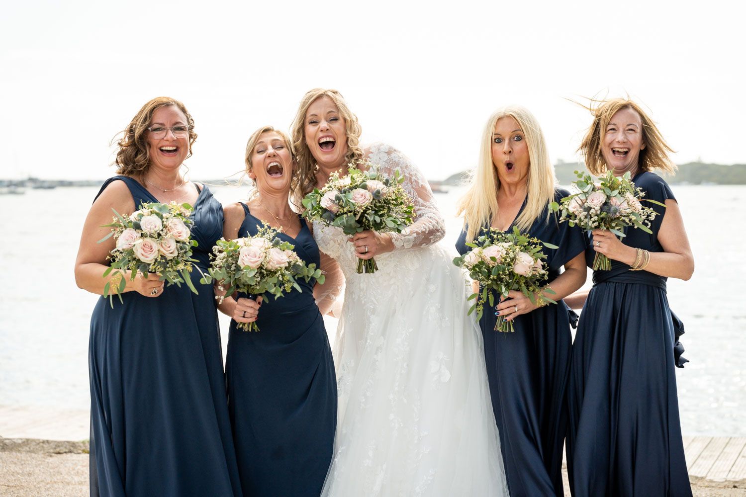 A wedding party in navy blue dresses and white gown stand laughing joyfully by the waterfront holding floral bouquets.