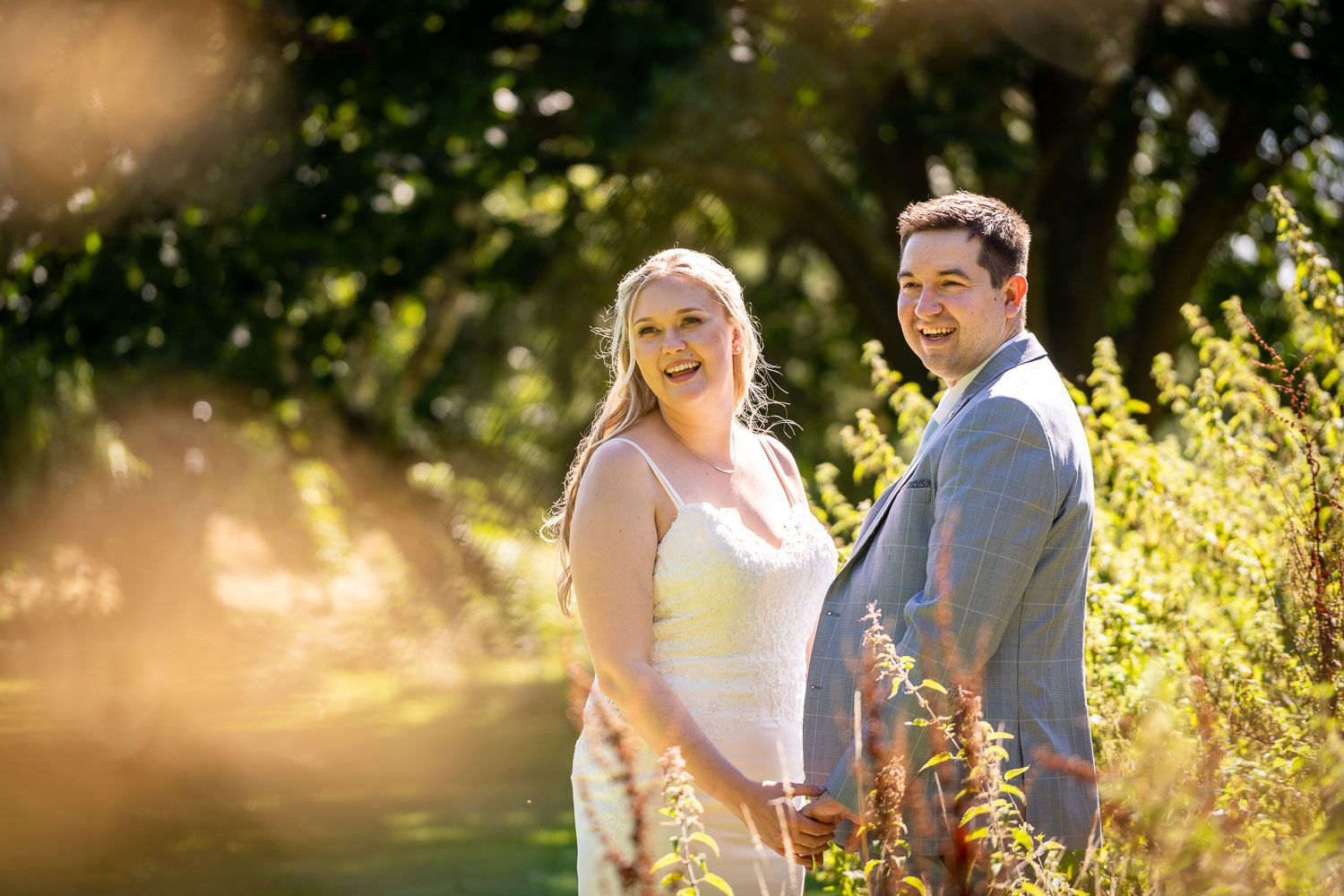 A couple stands in a sunlit meadow during golden hour, with warm light filtering through trees and tall grass.