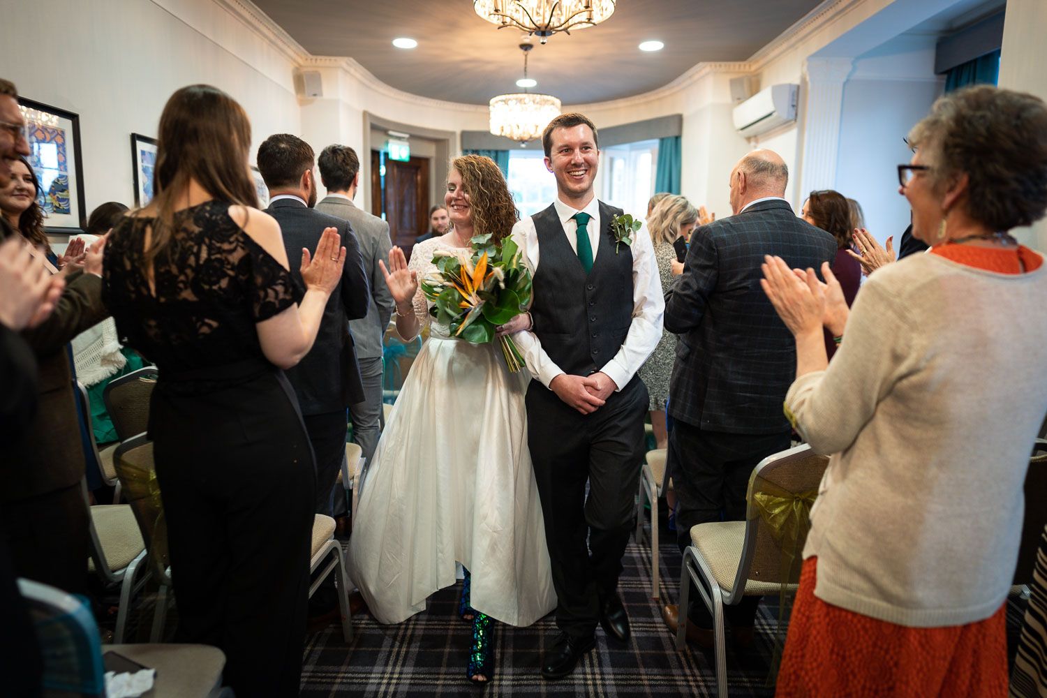 A couple stands together at a wedding reception as guests applaud around them in an elegant venue with chandelier lighting.