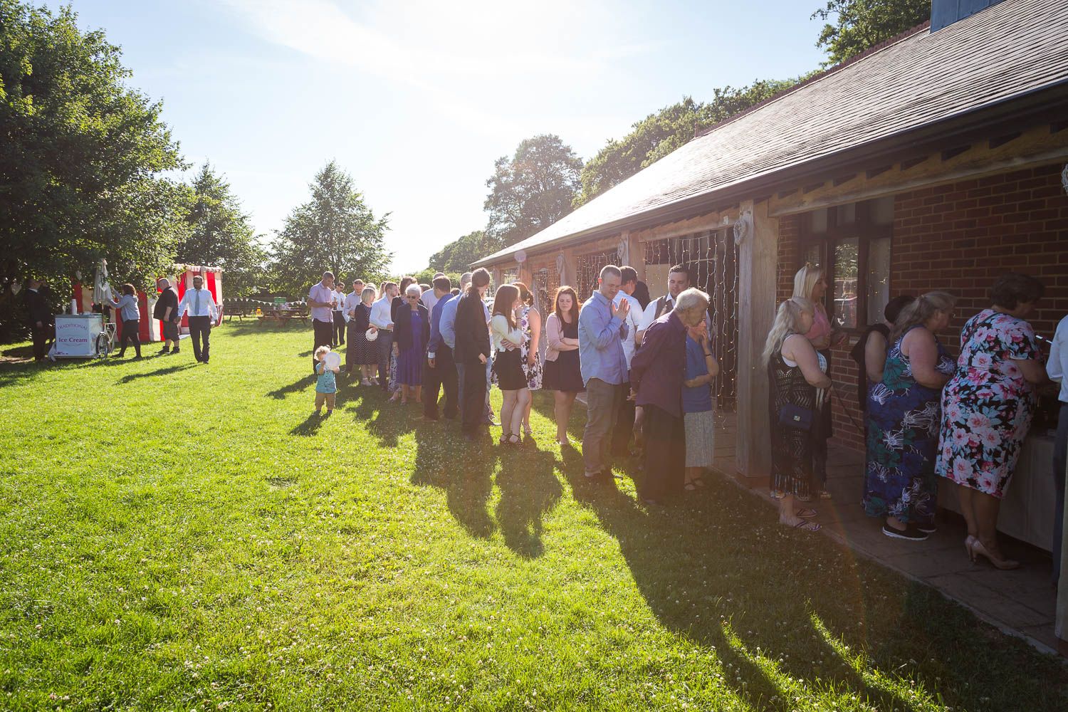 People gather at an outdoor event venue with a brick building and green lawn on a sunny summer afternoon.