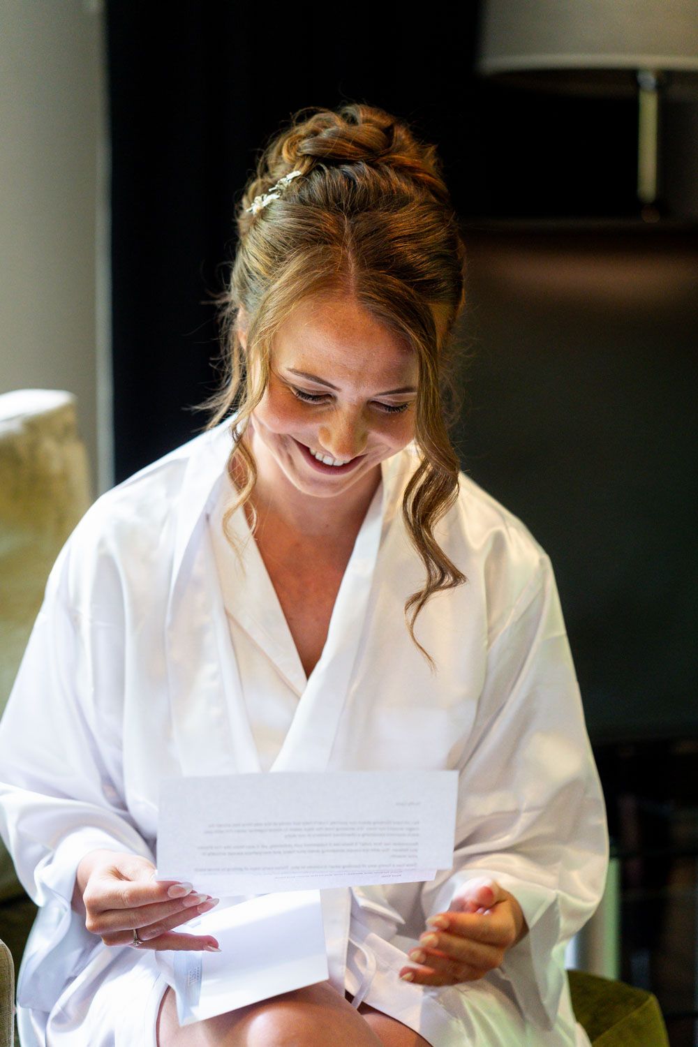 A joyful person in a white robe reads a letter while smiling warmly during wedding preparations.
