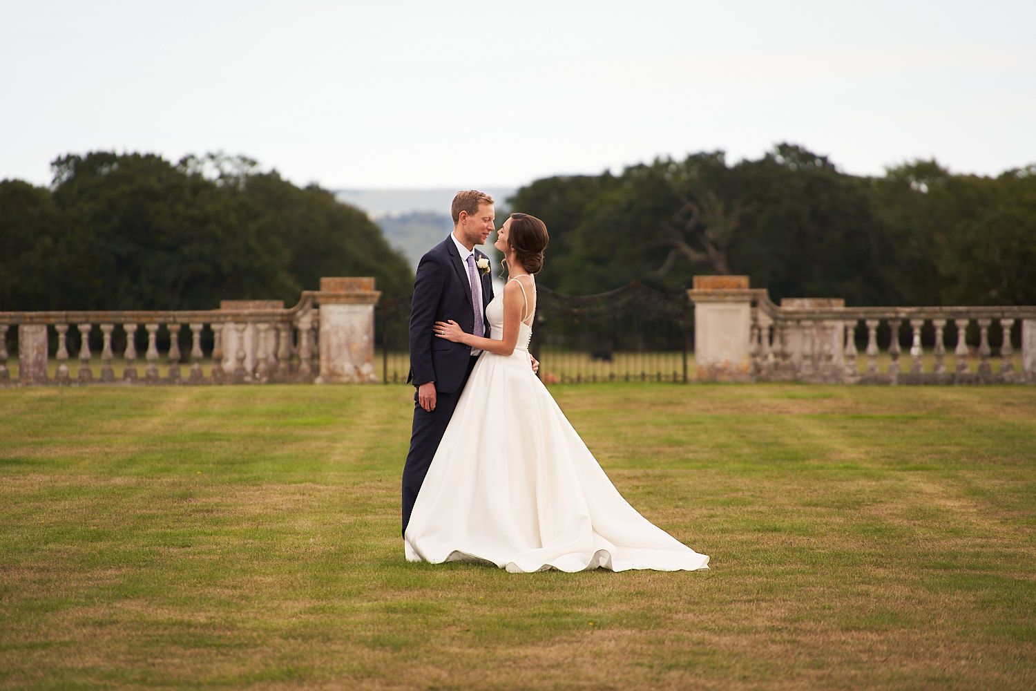 A bride and groom share a romantic moment on the lawn of an elegant estate with stone balustrades at sunset.