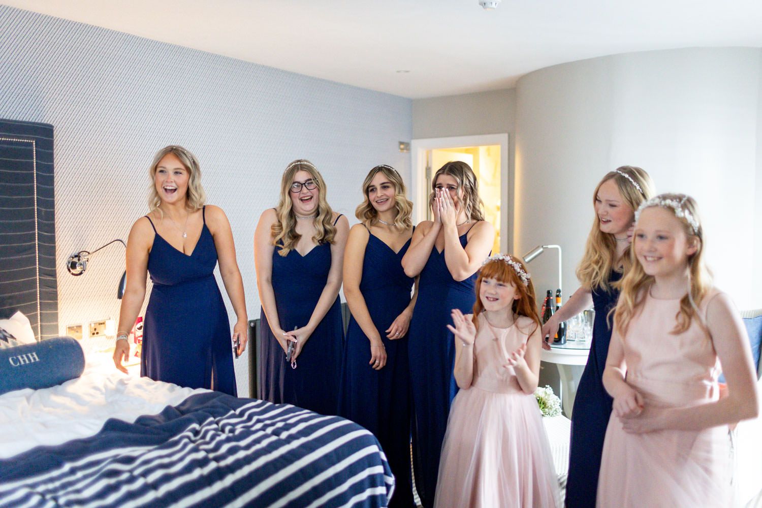 A group in navy blue bridesmaid dresses and pink flower girl dresses gather together in a bright hotel room.