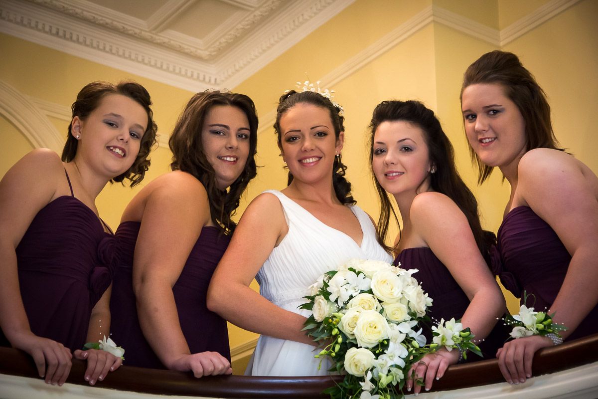 Wedding party in purple bridesmaid dresses poses on staircase with bride in white holding cream bouquet.