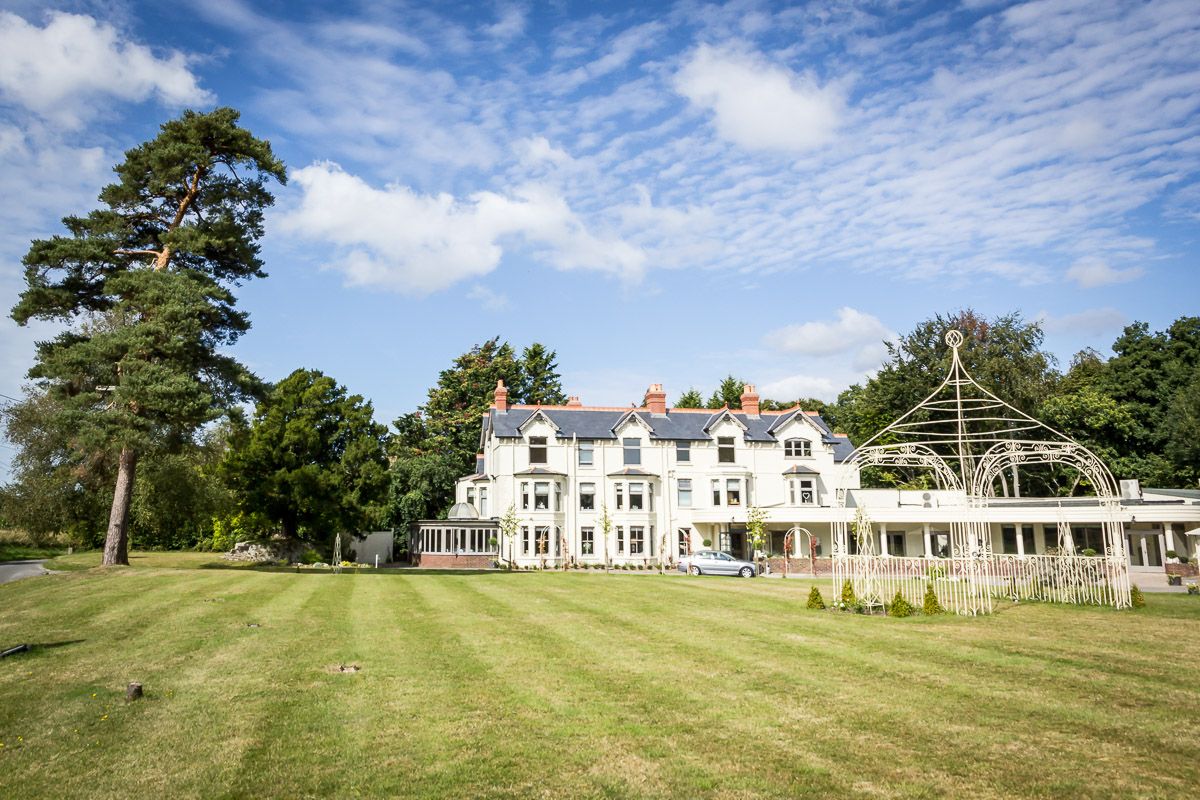 A grand white Victorian mansion with large windows sits on a manicured lawn under a bright blue summer sky.