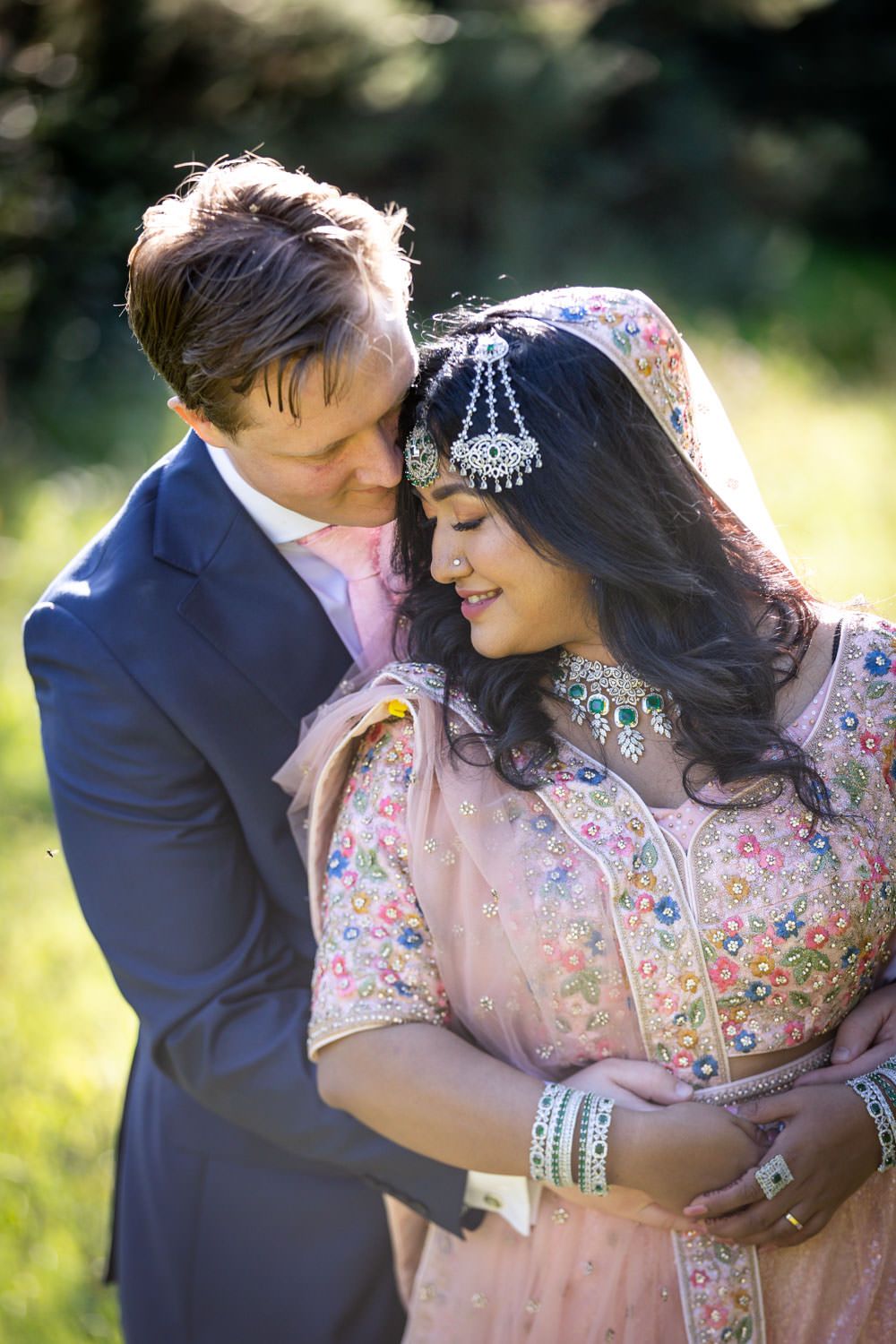 Groom in blue suit embracing bride wearing a beautiful pink embroidered Indian wedding outfit.