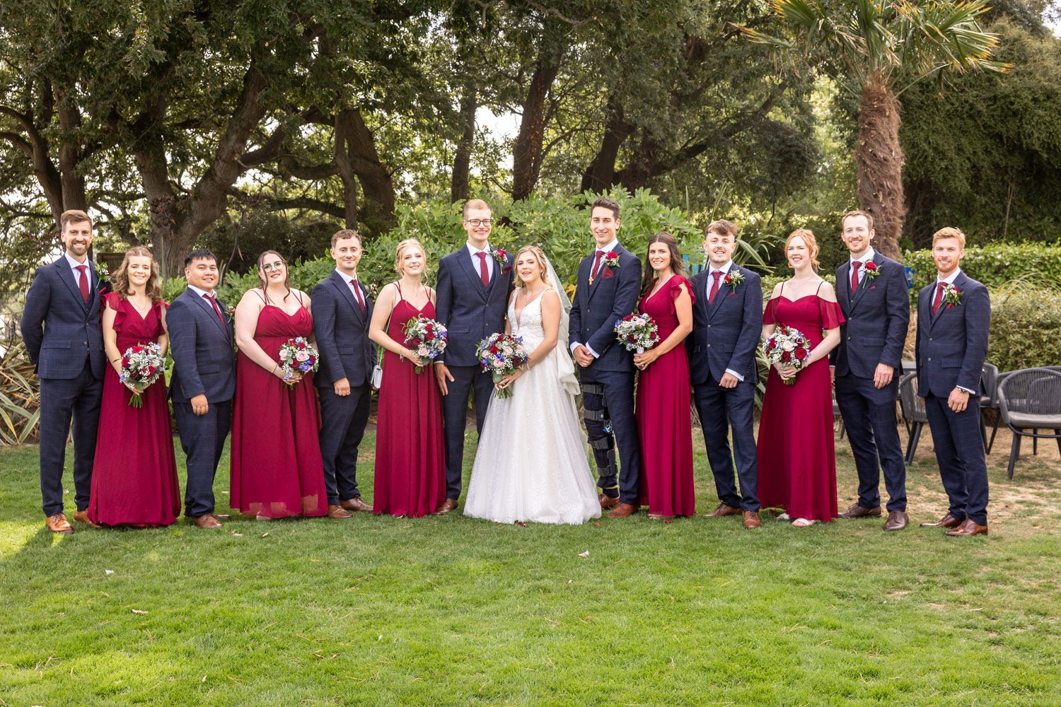 Wedding party poses outdoors with bridesmaids in red gowns and groomsmen in navy suits flanking the bride and groom.