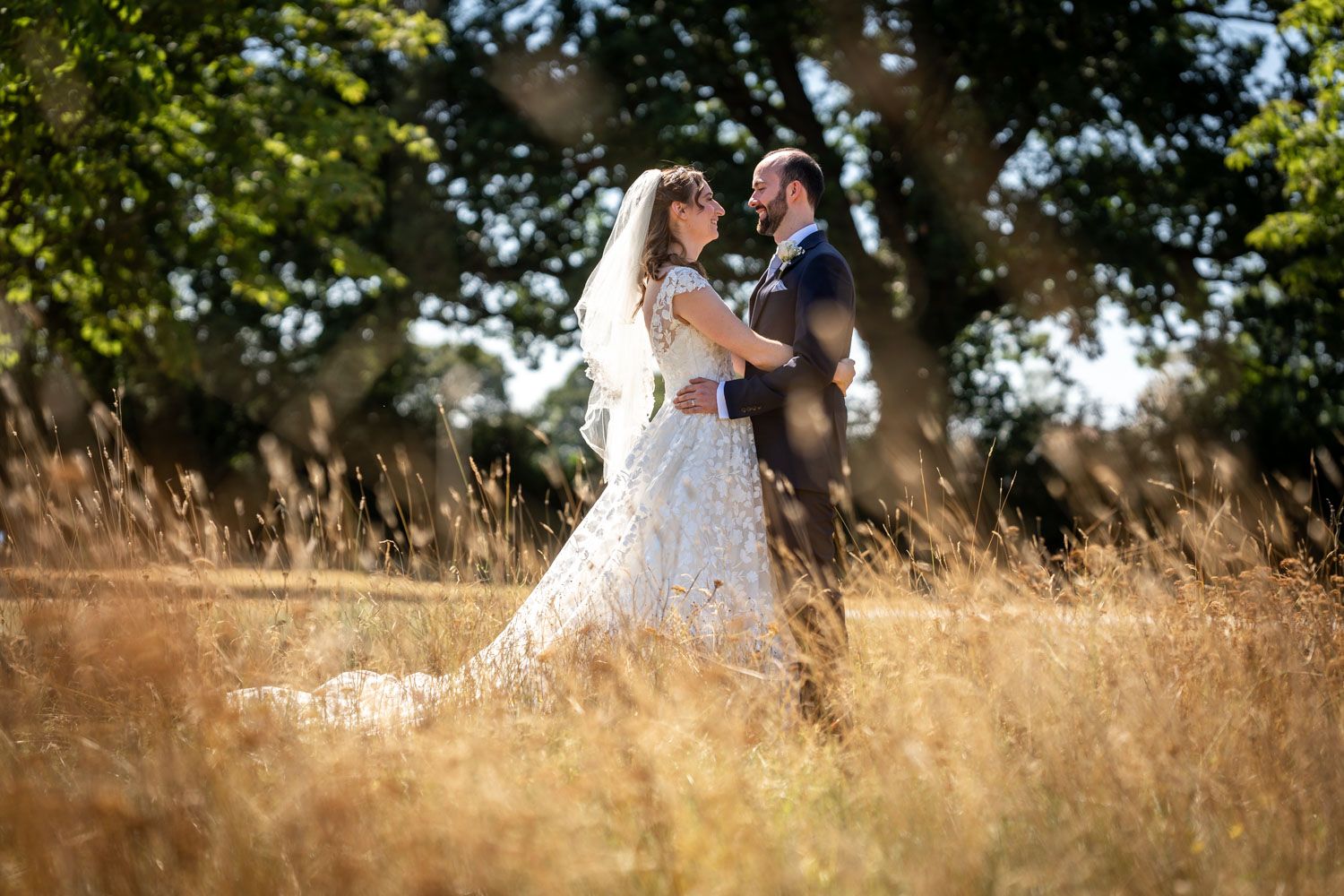 Bride and groom embrace romantically in a golden sunlit field with tall grass and trees behind.
