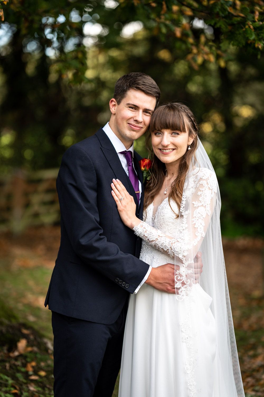 A bride in a long-sleeved lace wedding dress and veil poses with her groom in a navy suit outdoors in an autumn setting.