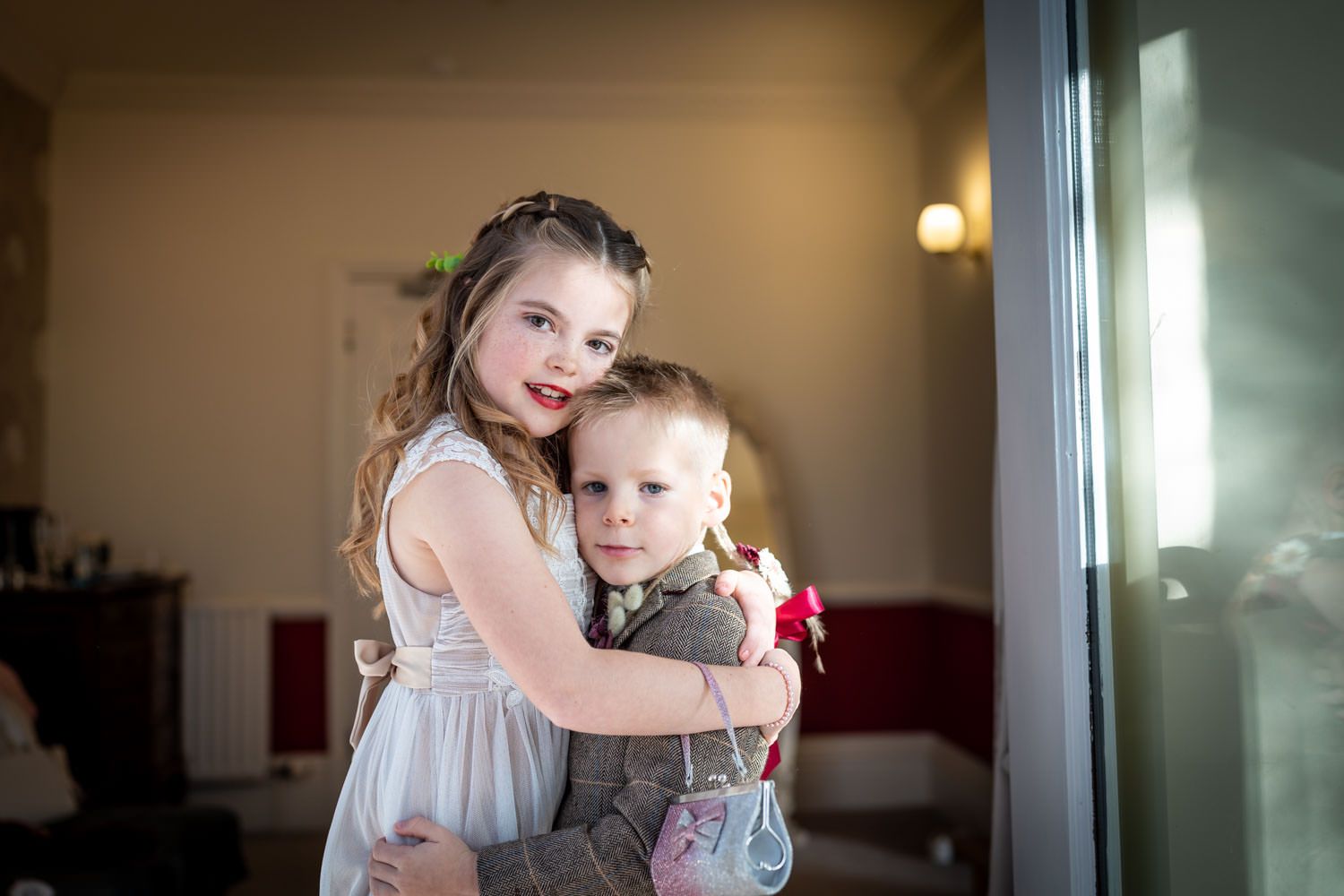 Two young children in formal attire embrace during a special occasion indoors.