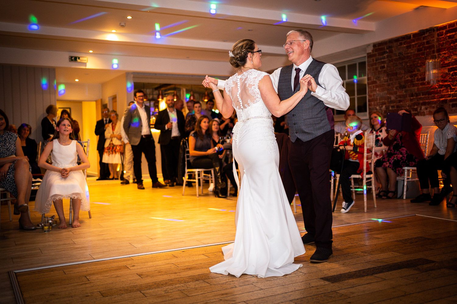 Wedding couple shares their first dance on a rustic wooden dance floor as guests watch in warm lighting.