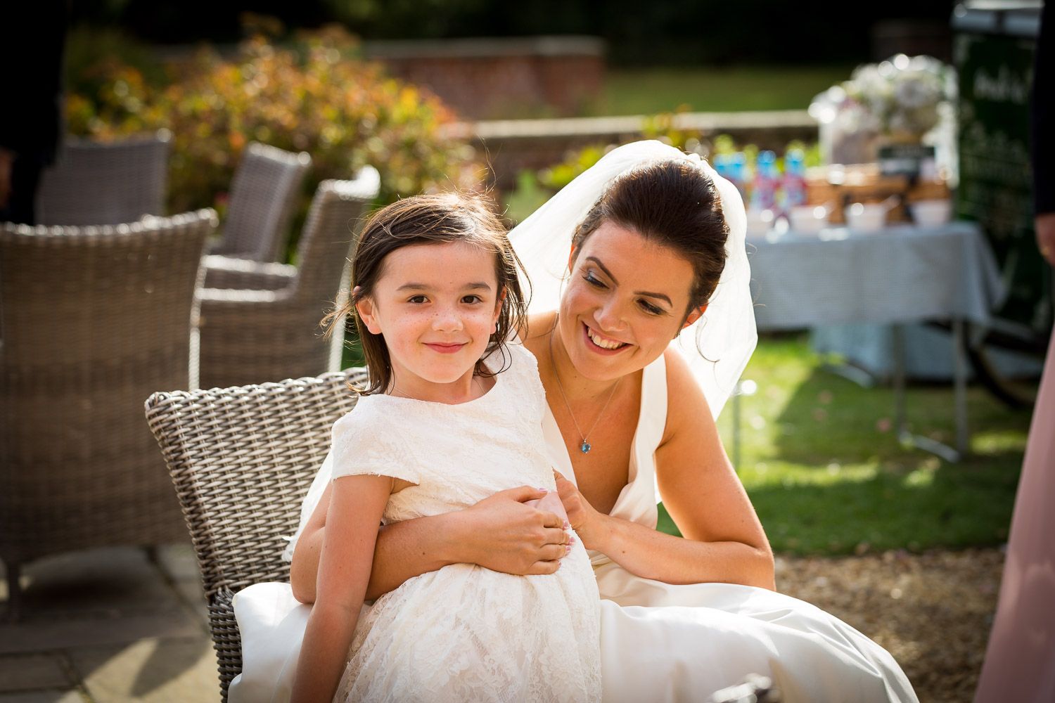 Two people in elegant white attire share a tender moment while seated on wicker patio furniture in a sunny garden setting.