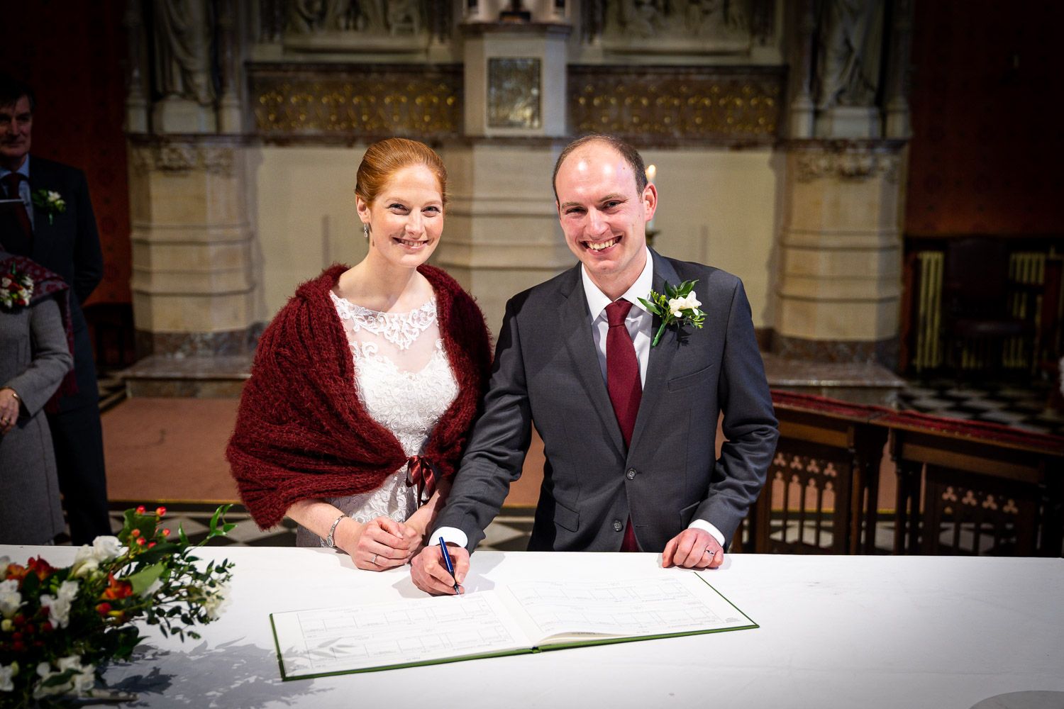 A couple signs a registry book during a church wedding ceremony, with the bride wearing a burgundy wrap shawl.
