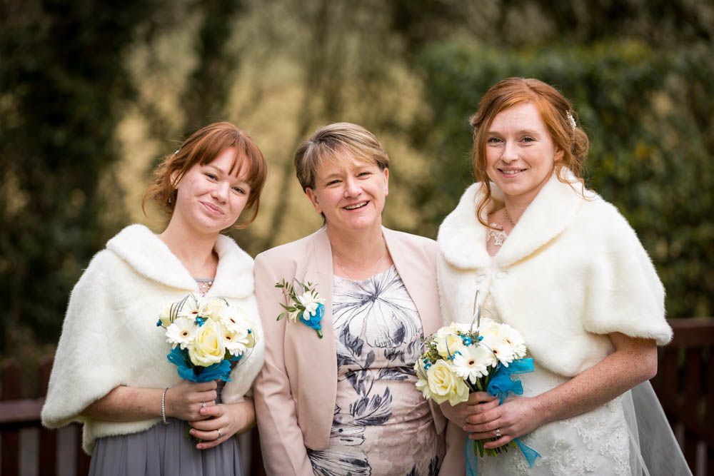 Three women in white shawls holding blue and white floral bouquets pose together outdoors.