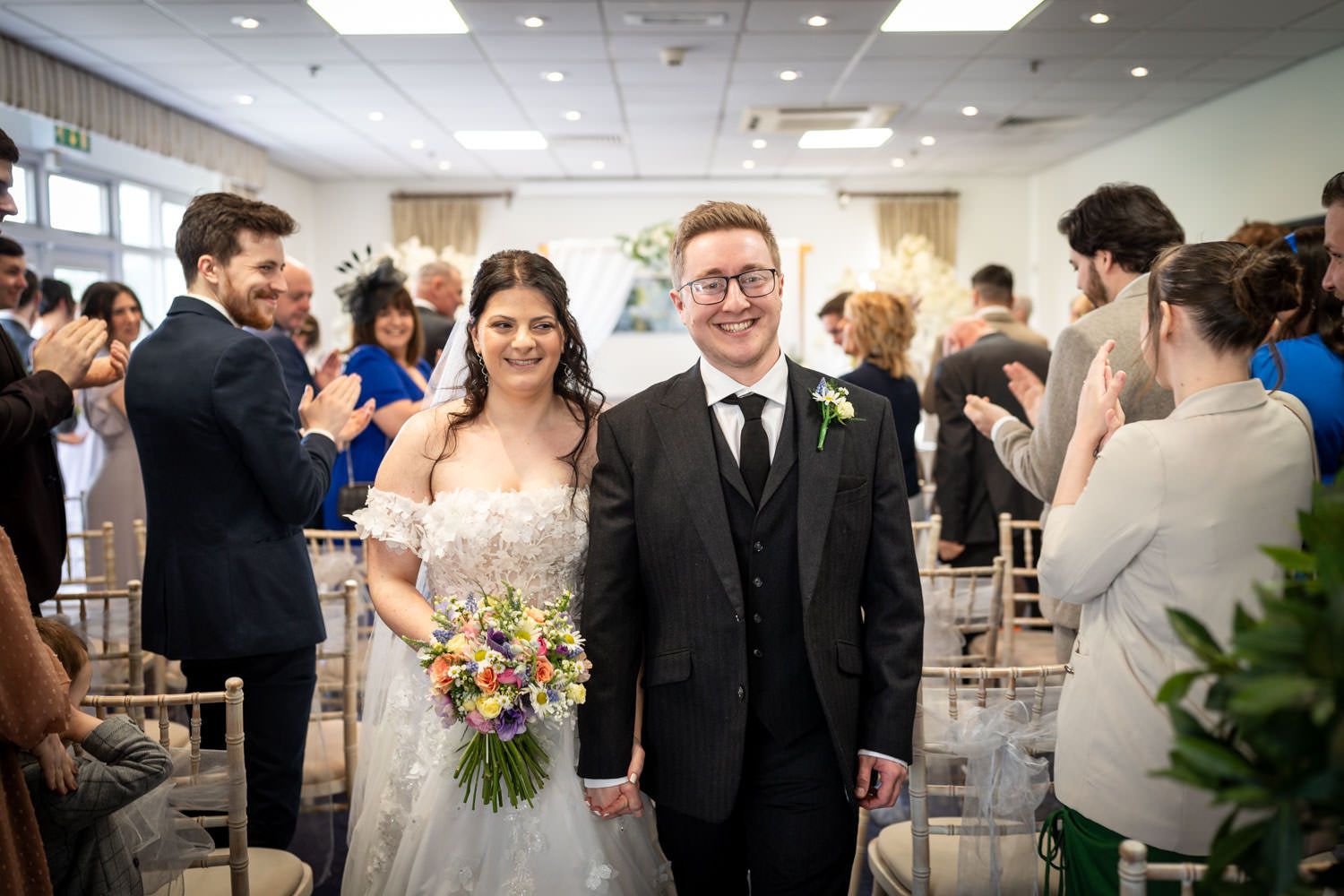 A newlywed couple beams with joy as they walk down the aisle at their indoor wedding ceremony.
